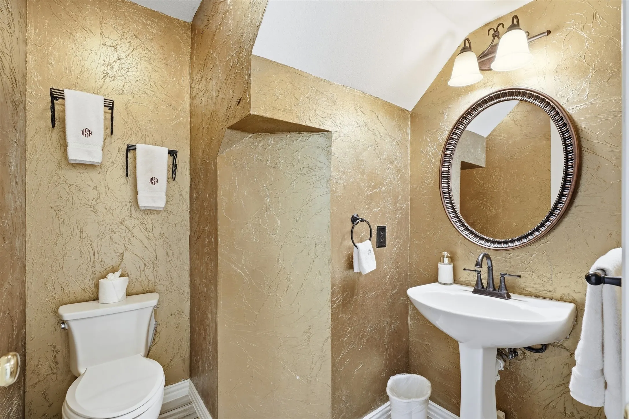 Bathroom featuring textured gold-toned walls, a white pedestal sink with a dark bronze faucet, and a round framed mirror