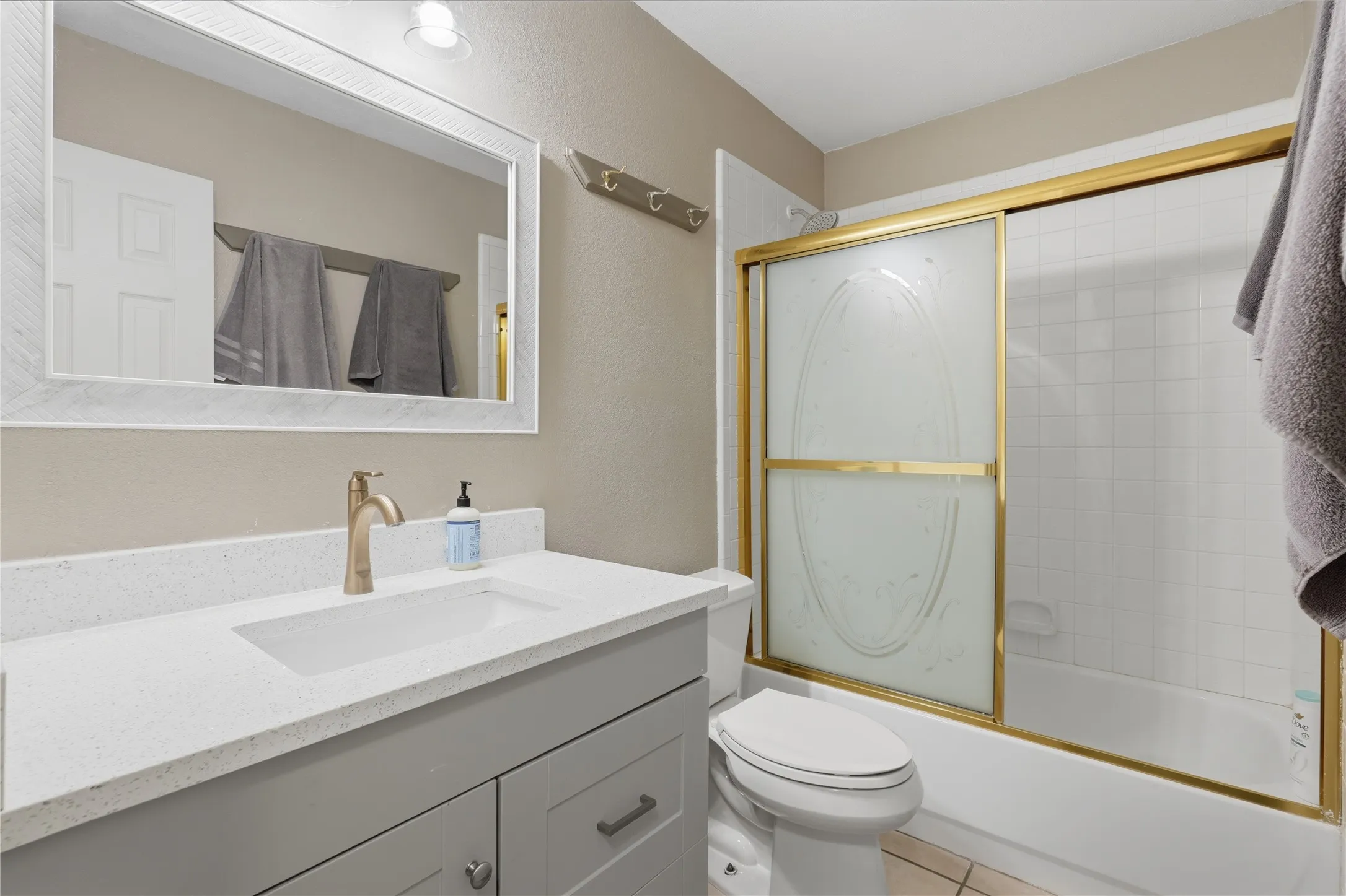 Bathroom featuring a light gray vanity with a quartz countertop and integrated sink, brushed gold-finish faucet, white-framed mirror, and a shower-tub combination with a gold-framed sliding door