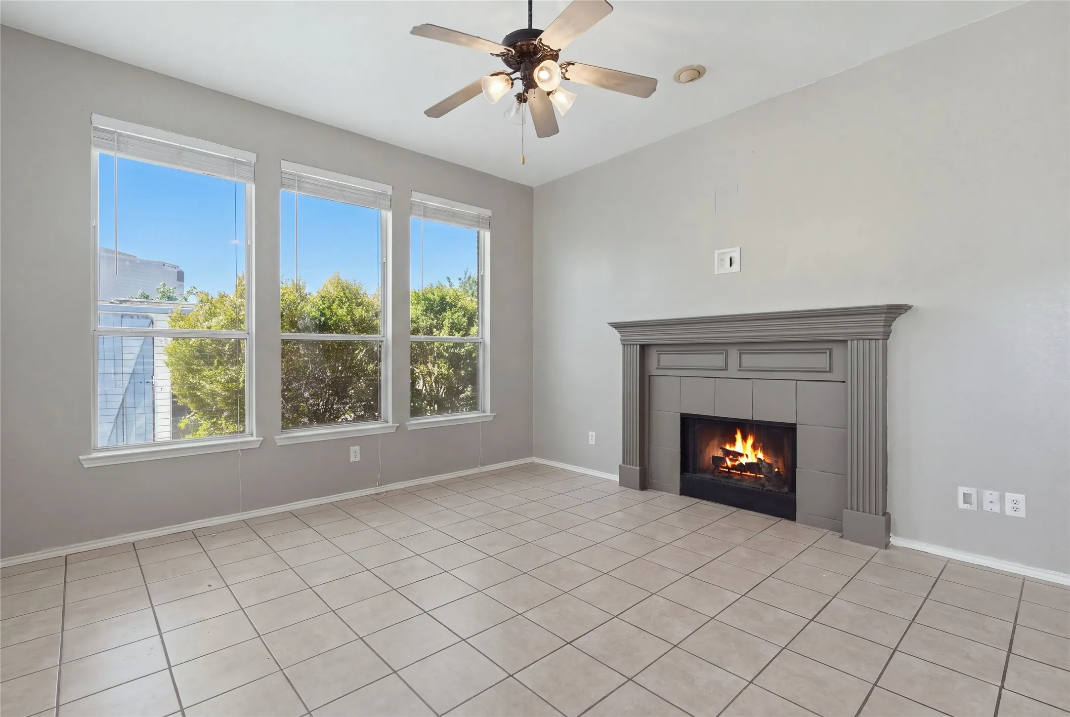 Spacious room featuring three large windows, a ceiling fan with integrated lighting, a fireplace with a decorative mantel, and tile flooring