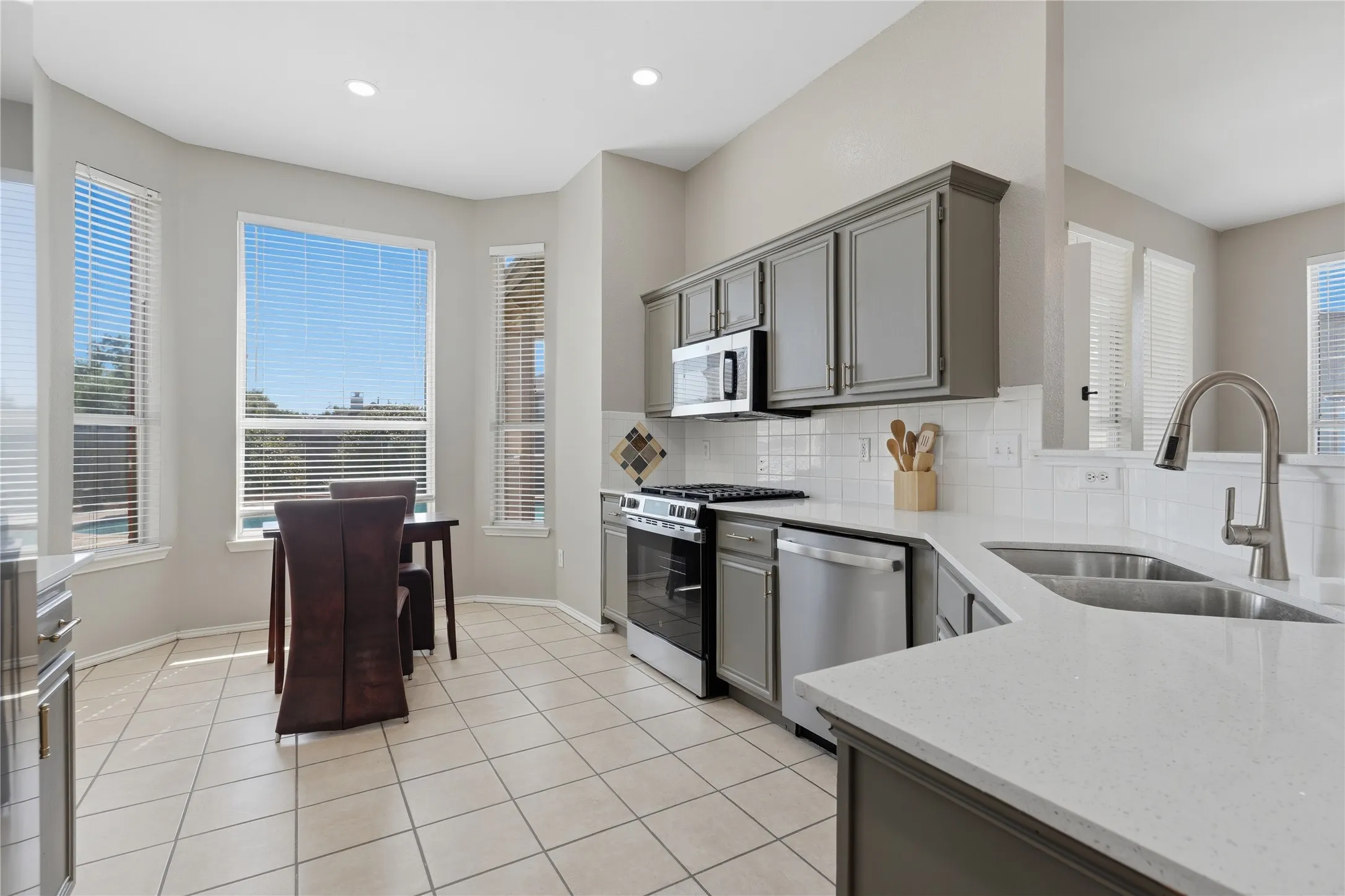 Kitchen featuring gray cabinetry, stainless steel appliances, white tile backsplash, quartz countertops, and a dual basin sink
