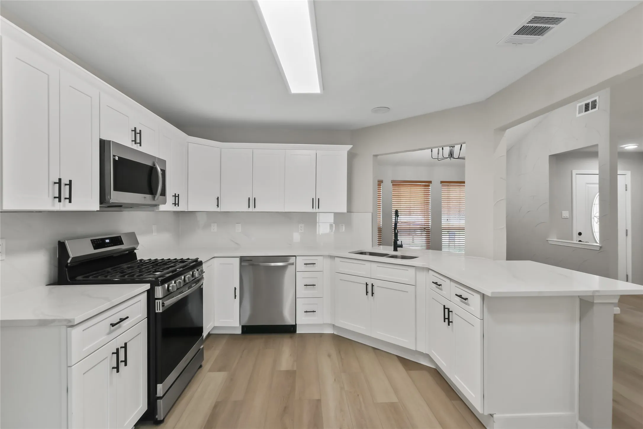 Modern kitchen featuring white shaker-style cabinetry with black hardware, stainless steel appliances including a gas range and built-in microwave, white subway tile backsplash, white quartz countertops, a large island with seating, and wood-finish flooring throughout the open layout