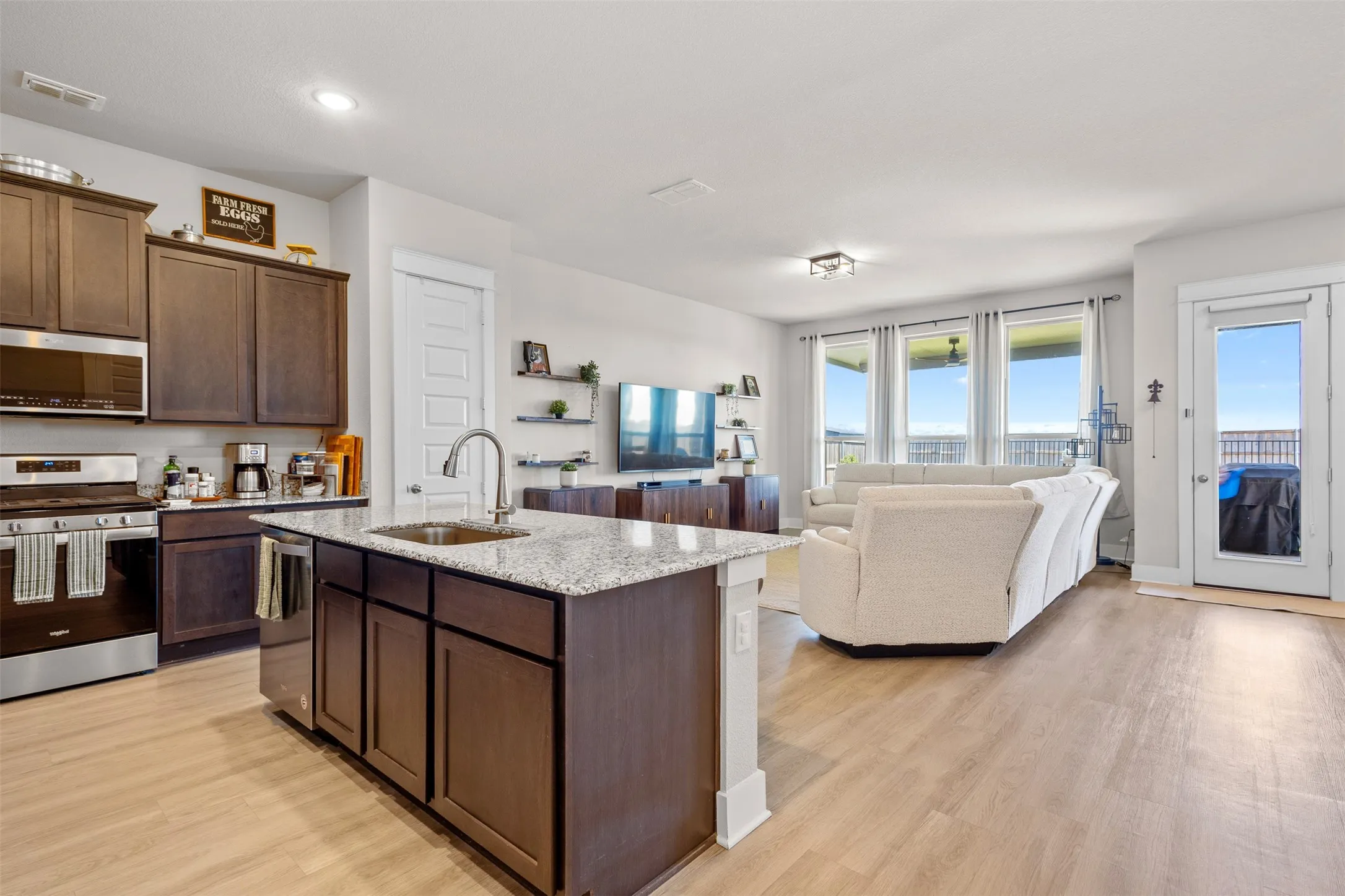 Open-concept interior featuring light wood-finish flooring, a kitchen island with integrated sink, stainless steel appliances, and a wall of windows