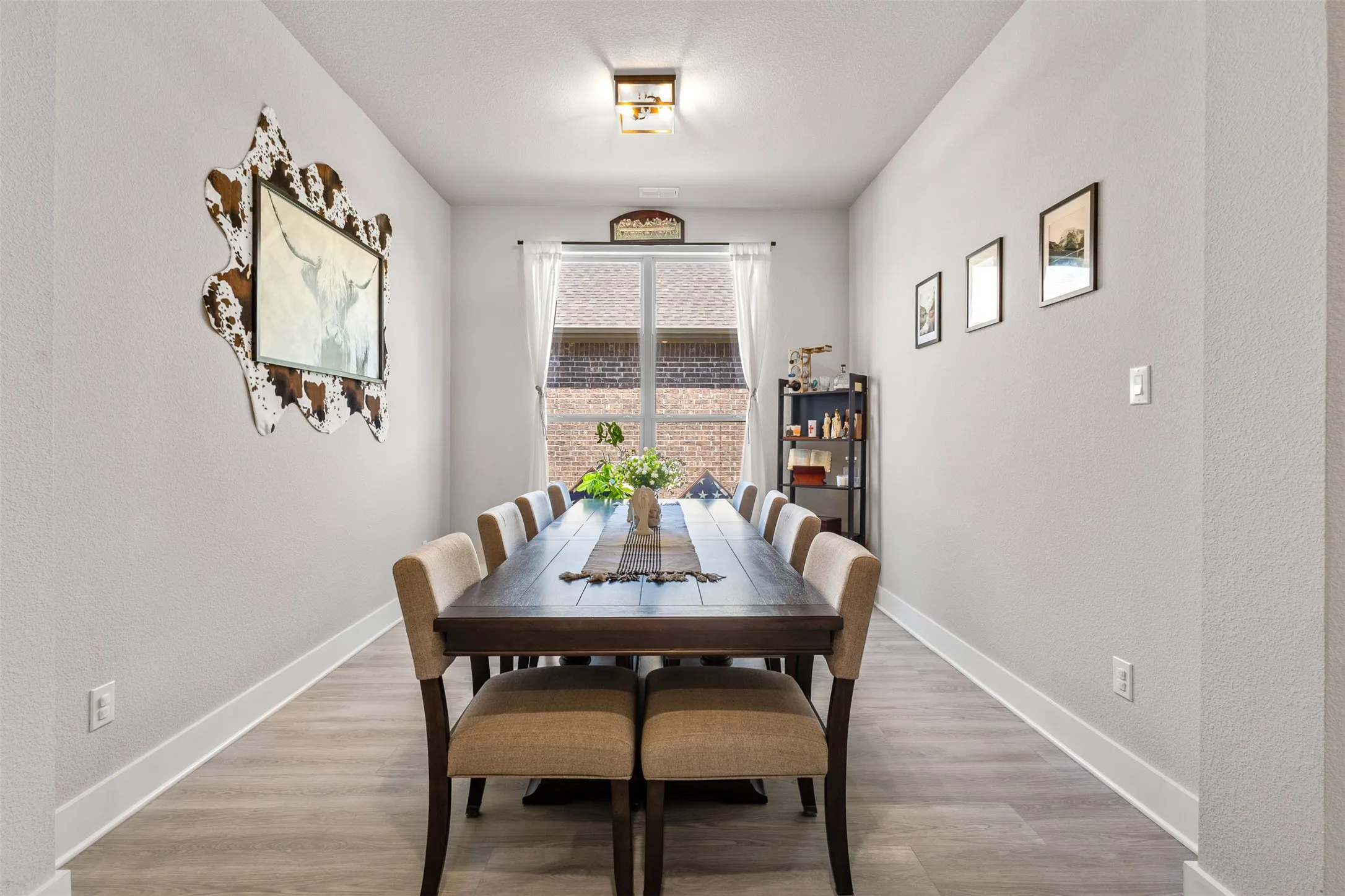 Long dining space with wood-finish flooring, a contemporary ceiling light fixture, and a window with white curtains