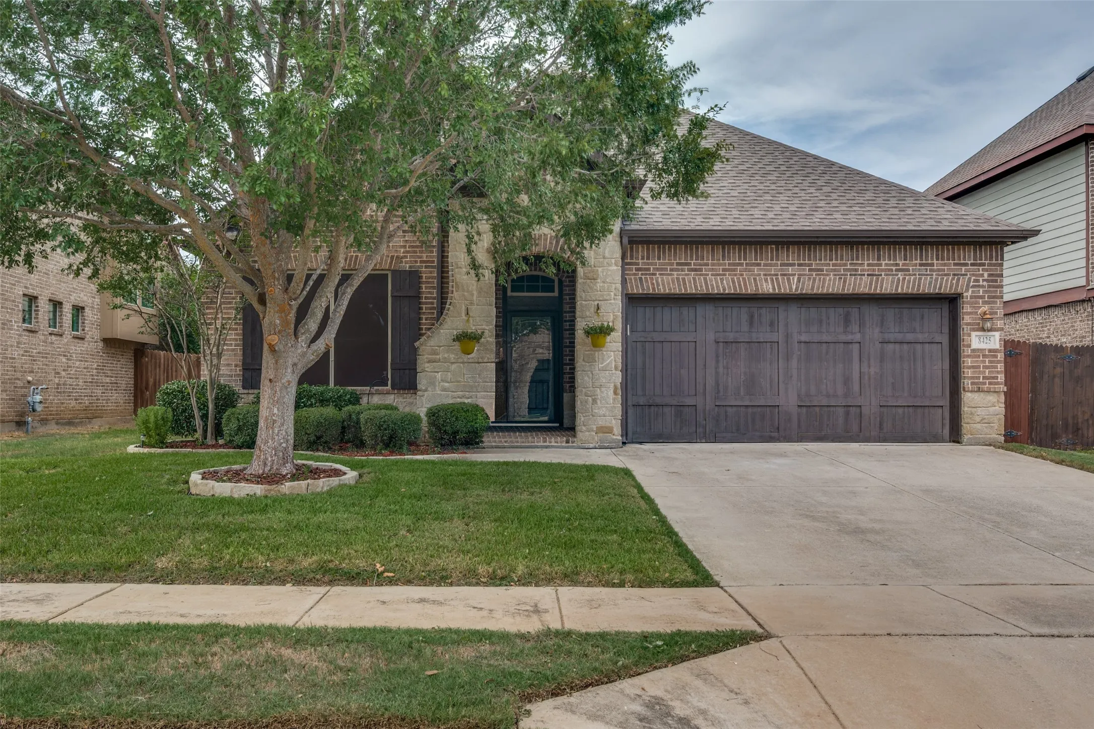 Brick and stone facade, two-car garage with wood-finish doors, mature front yard tree, concrete driveway, and front walk