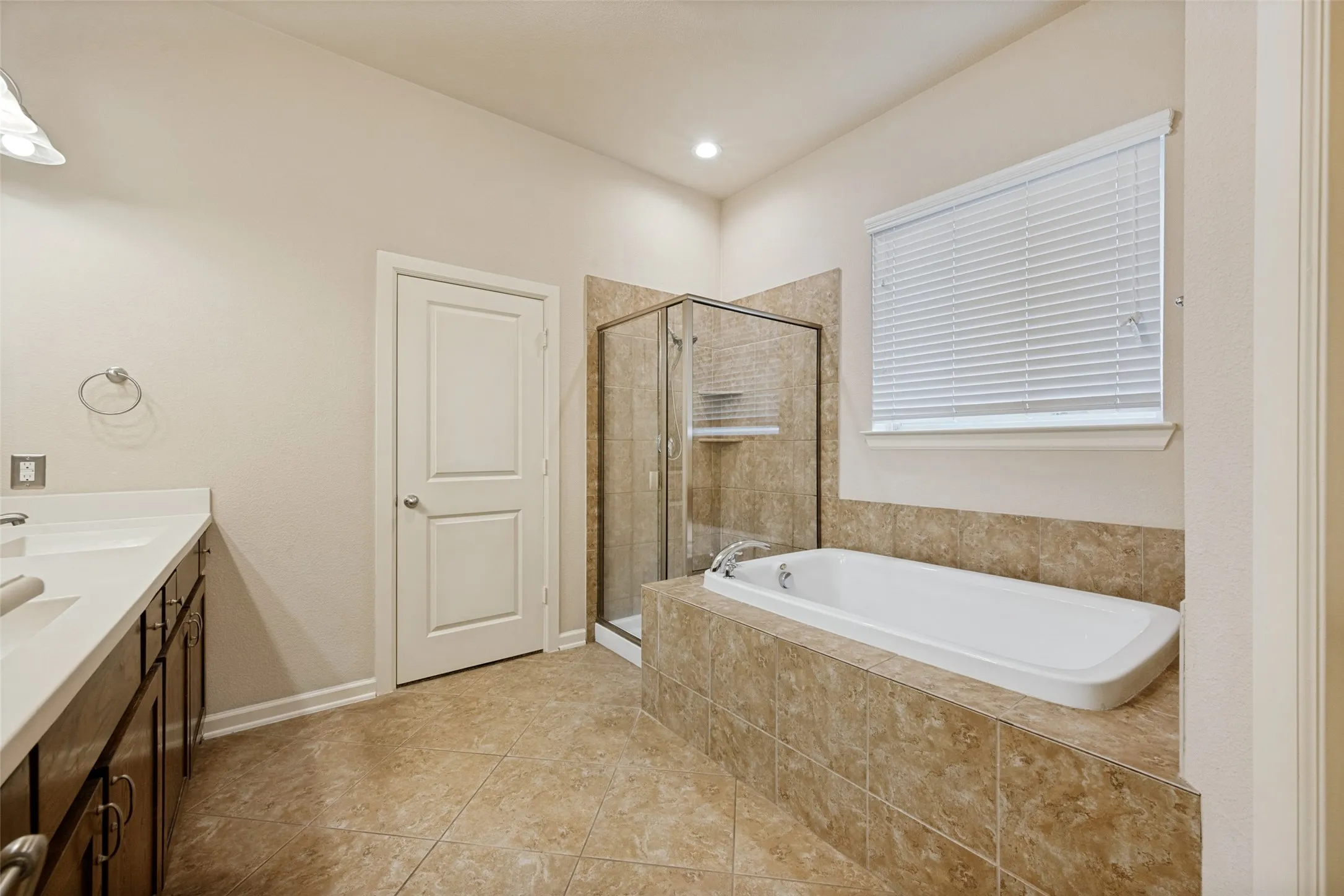 Dual vanity with white countertops, built-in soaking tub, tiled tub surround, glass-enclosed shower, and tile flooring