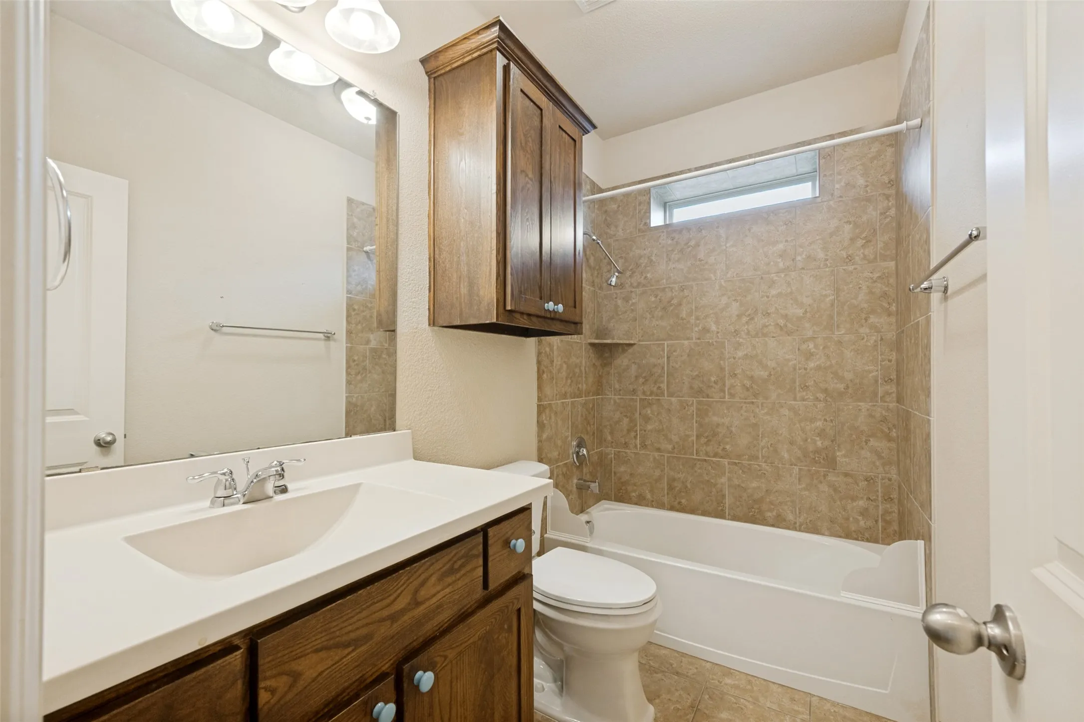 Bathroom featuring a single vanity with a white countertop, wood-finish cabinetry, an overhead wood-finish cabinet, a tiled shower/tub combination, and a window