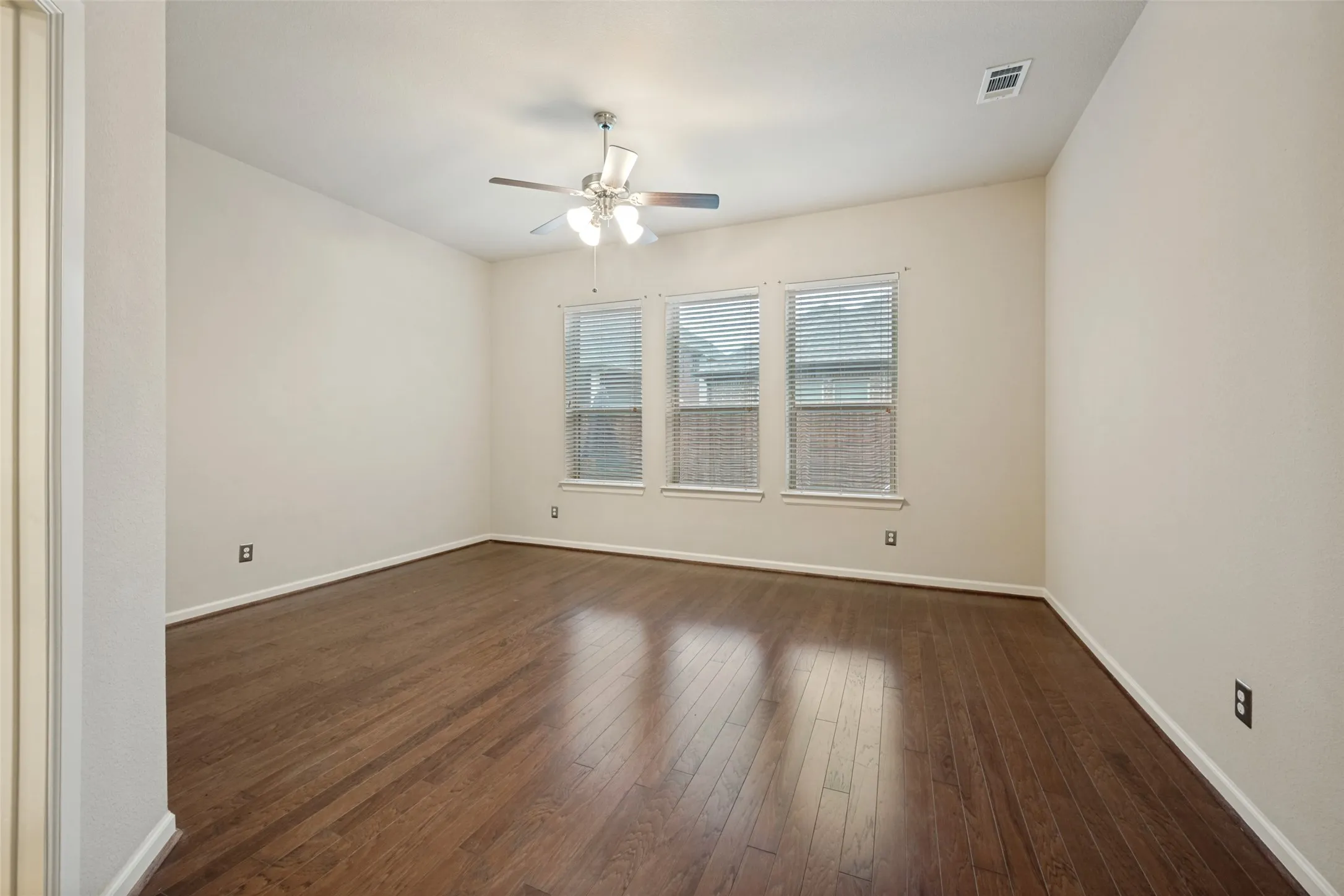 Hardwood flooring, three windows with blinds, ceiling fan, neutral wall color, and white trim