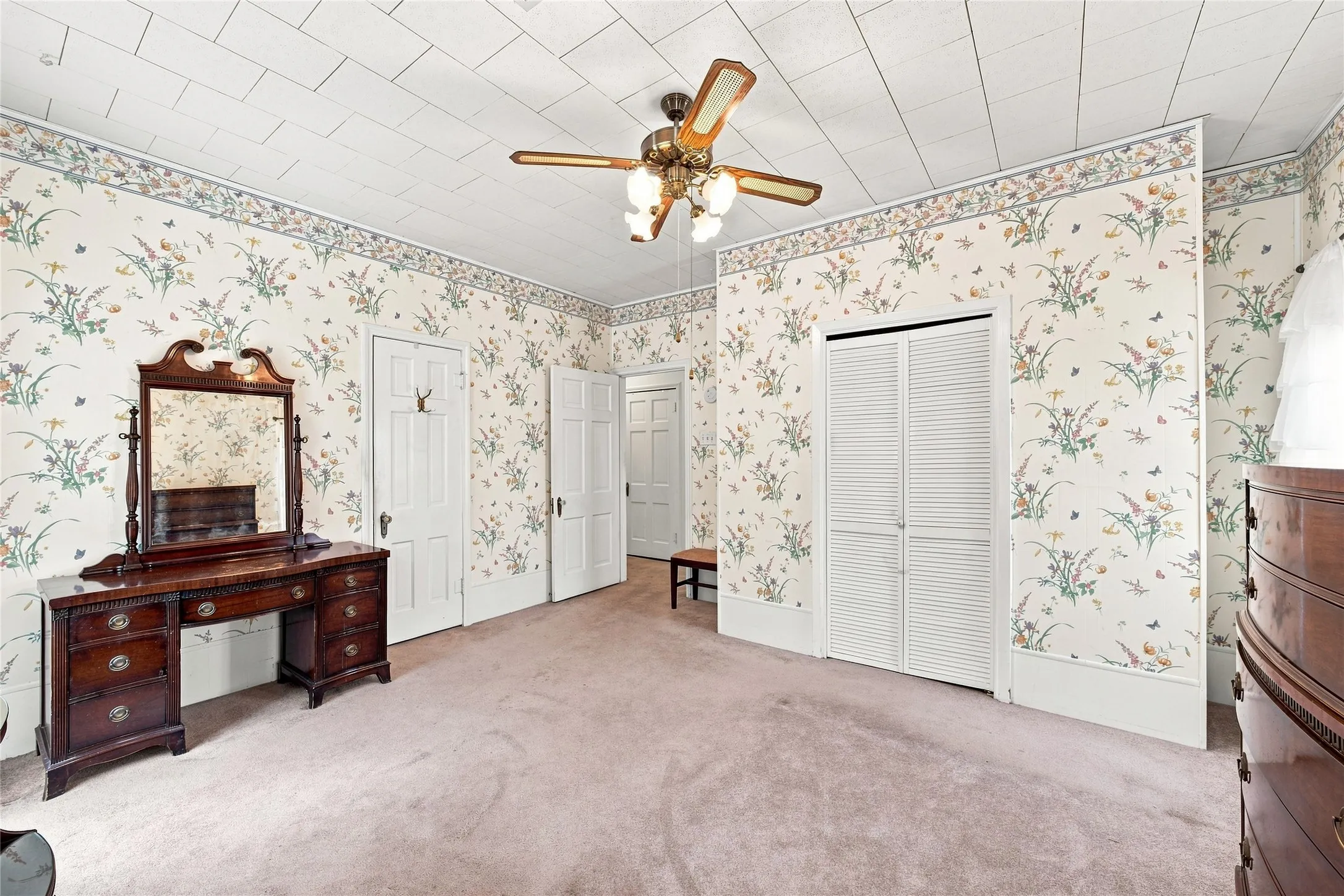 Carpeted flooring, patterned wallpaper, ceiling fan with integrated lighting, multiple white paneled doors, and white louvered closet doors