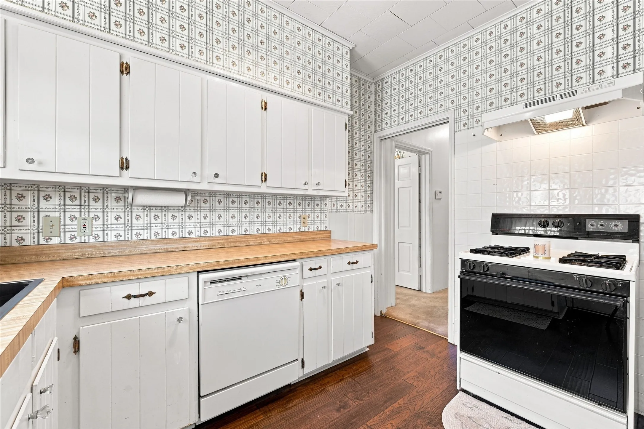White cabinetry, wood-finish countertops, white tiled backsplash, built-in dishwasher, and a white and black range with matching hood