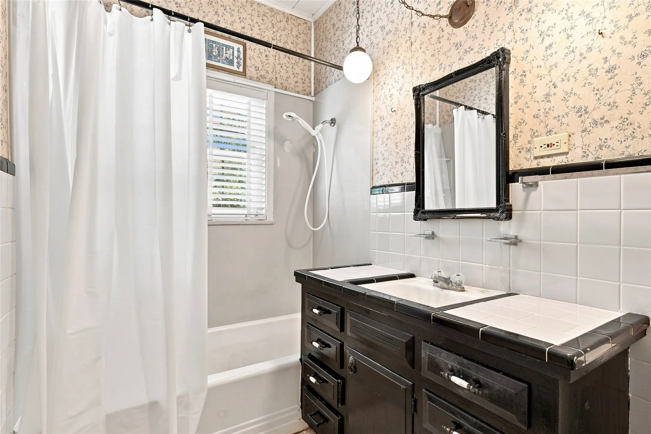 Window with blinds, white subway tile surround, dark wood-finish vanity with dual sinks, decorative framed mirror, and floral wallpaper
