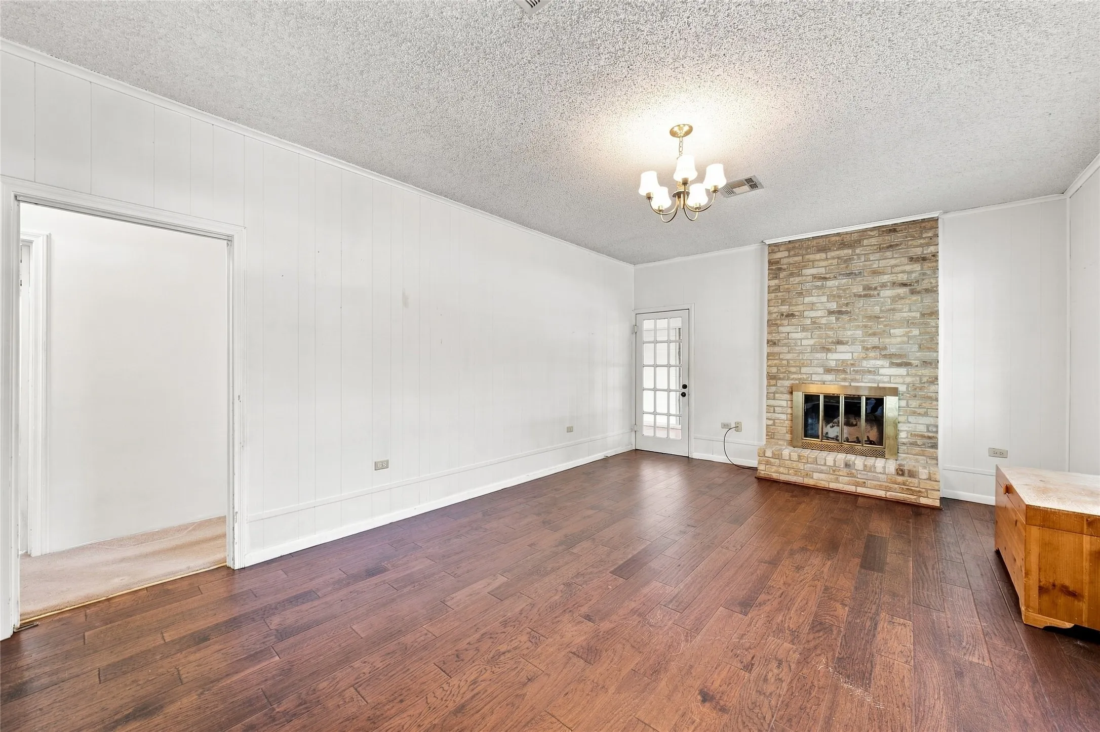 Spacious living room showcasing rich wood-finish flooring, white walls, and a brick fireplace with dark metal surround