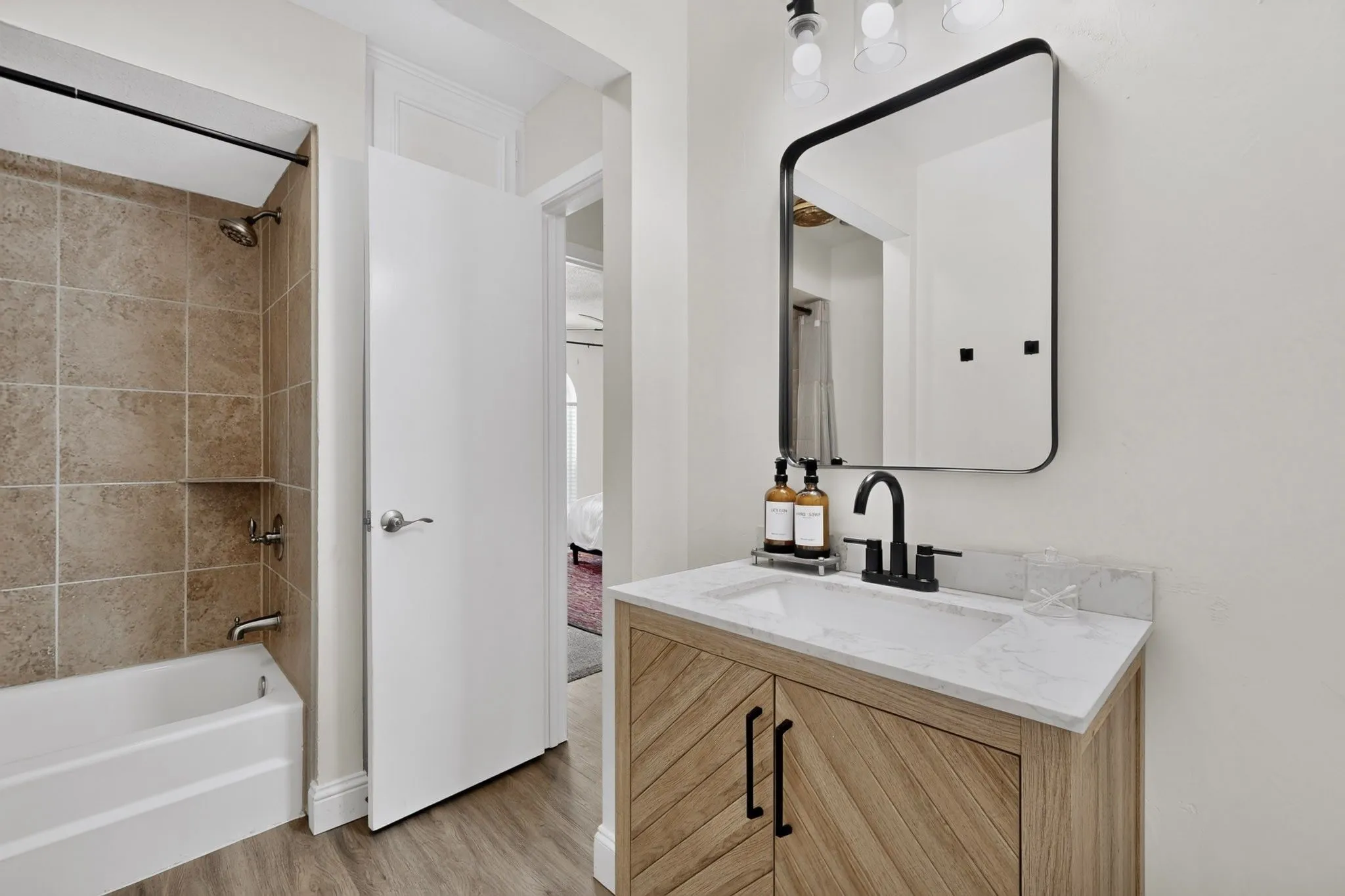 Wood-finish flooring, a vanity with a white countertop and integrated sink, a rectangular framed mirror, a tub/shower combination with tile surround, and a built-in shower niche