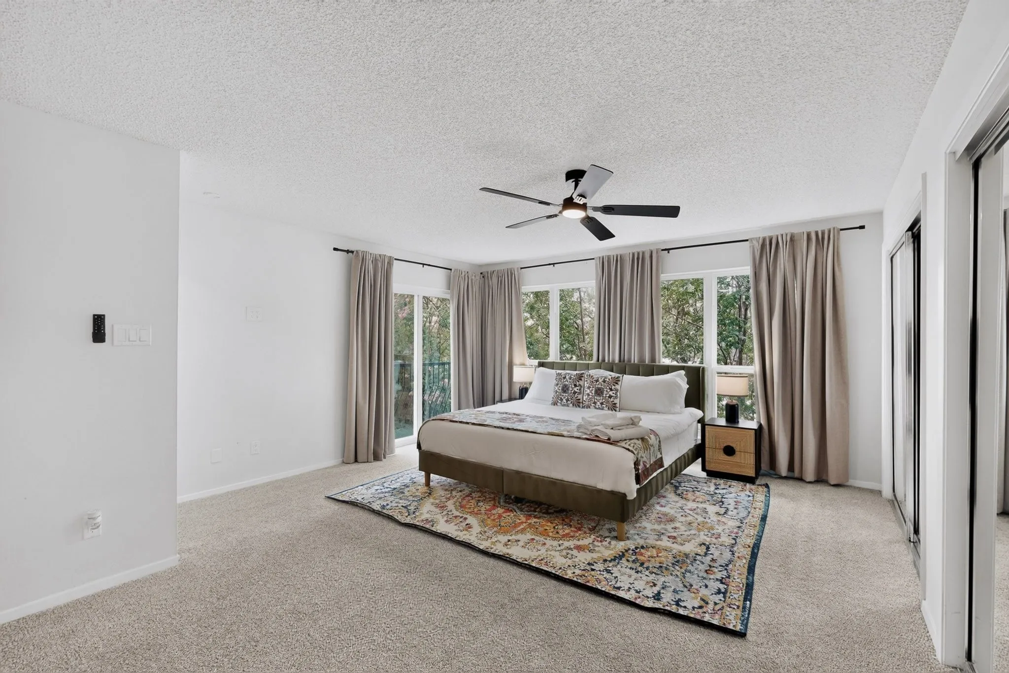 Spacious primary bedroom flooded with natural light through triple-pane windows, featuring neutral carpeting, ceiling fan with integrated lighting, and hardwood nightstands with integrated task lighting