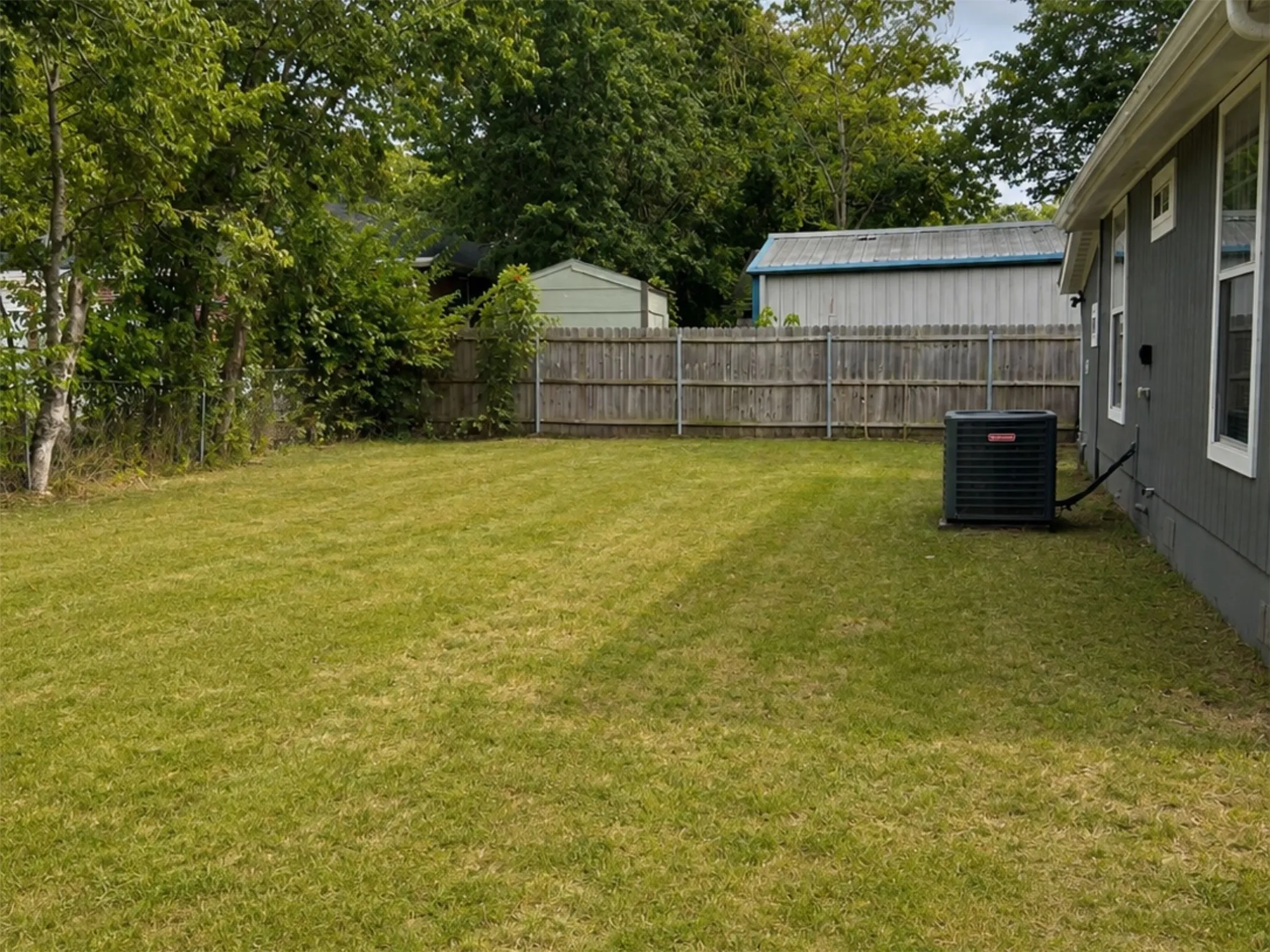 Expansive green lawn with a wooden privacy fence, accompanied by mature perimeter trees and side house paneling