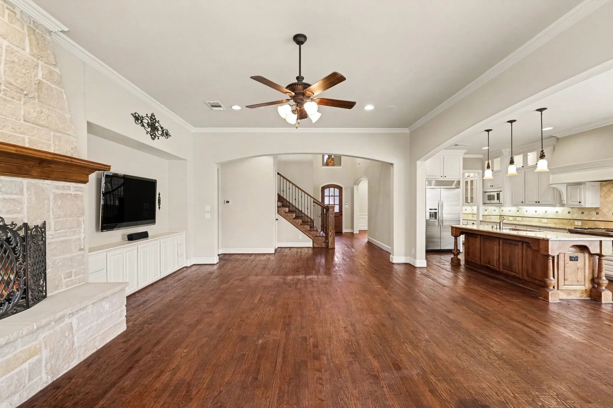 Hardwood flooring throughout, stone fireplace with custom mantel, built-in media cabinetry, open archway to kitchen, and a wooden staircase with iron balusters