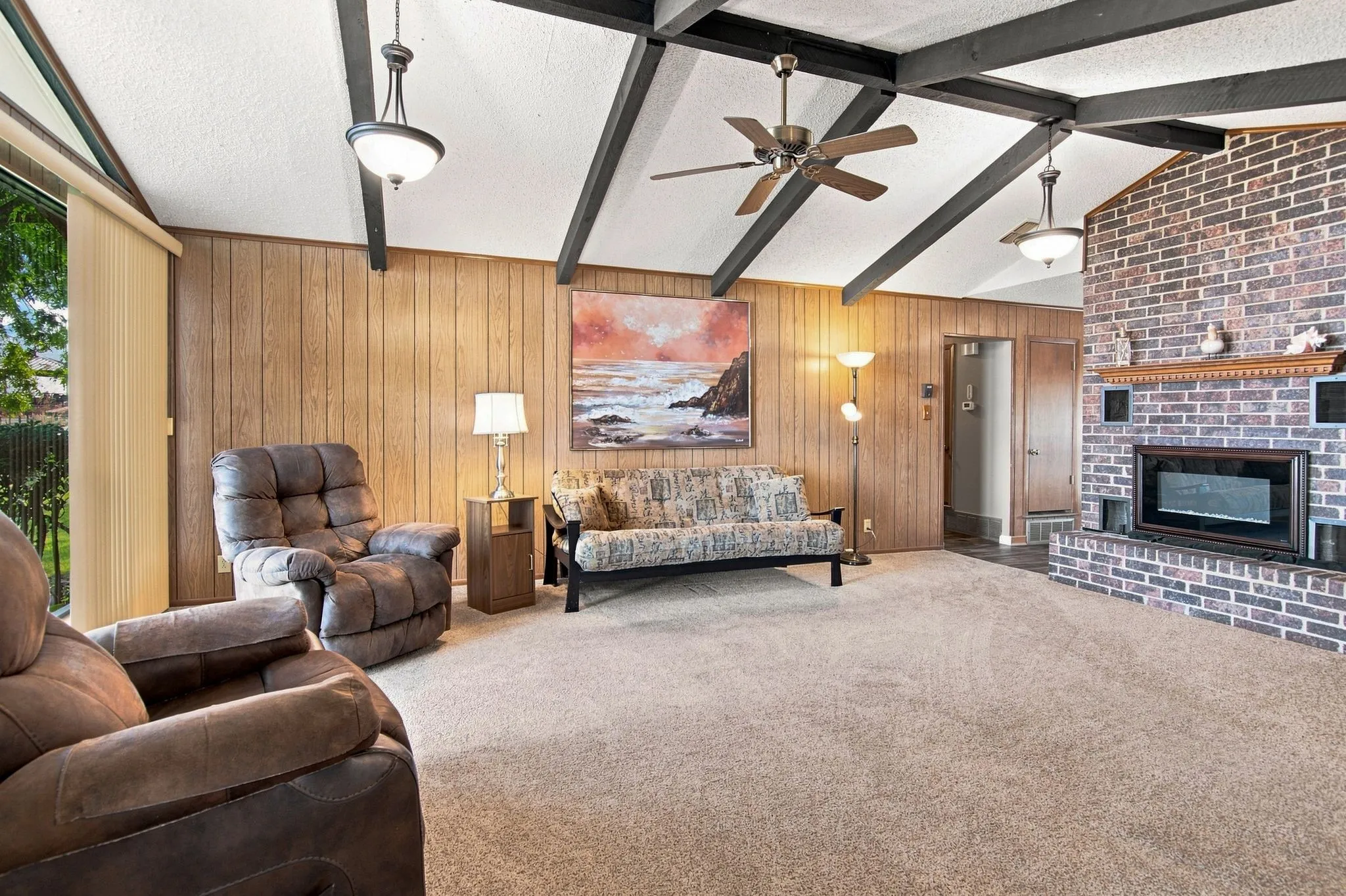 Vaulted ceiling with exposed beams, brick fireplace with a wood mantle, wood paneling, sliding glass door, and ceiling fan