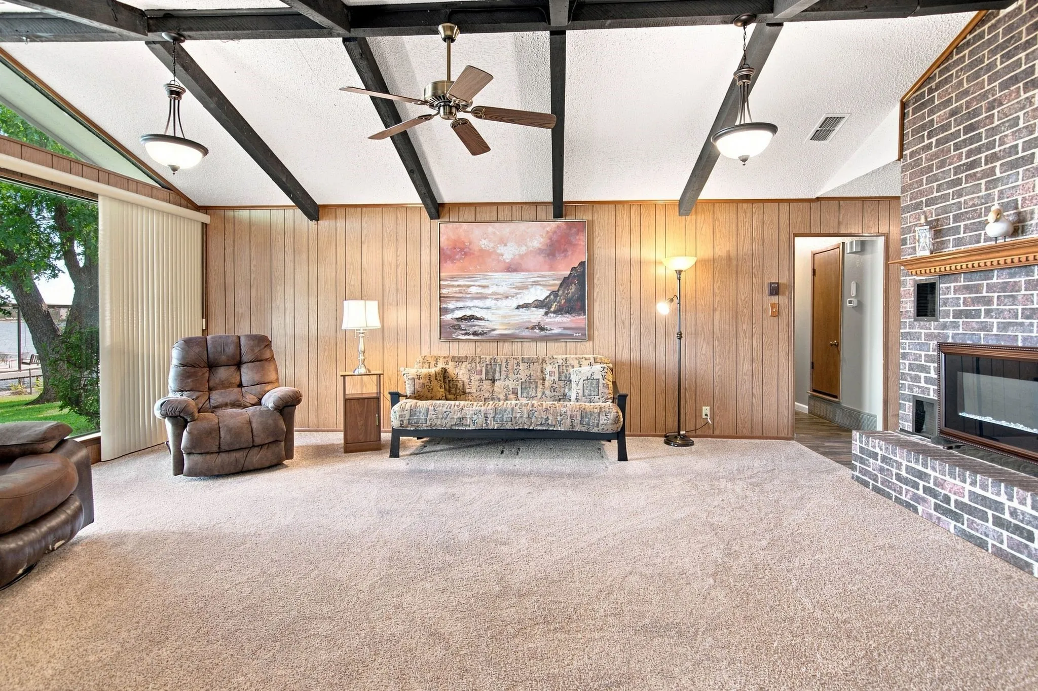 Vaulted ceiling with exposed beams, brick fireplace with a wood mantle, wood-paneled walls, and expansive windows