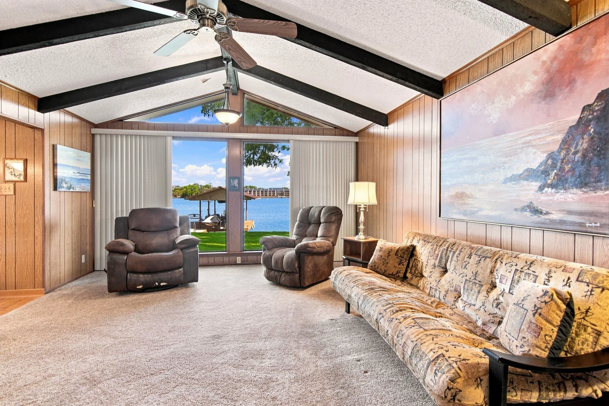 Vaulted ceiling with exposed beams, wood-paneled walls, carpeted flooring, and a ceiling fan with light fixture