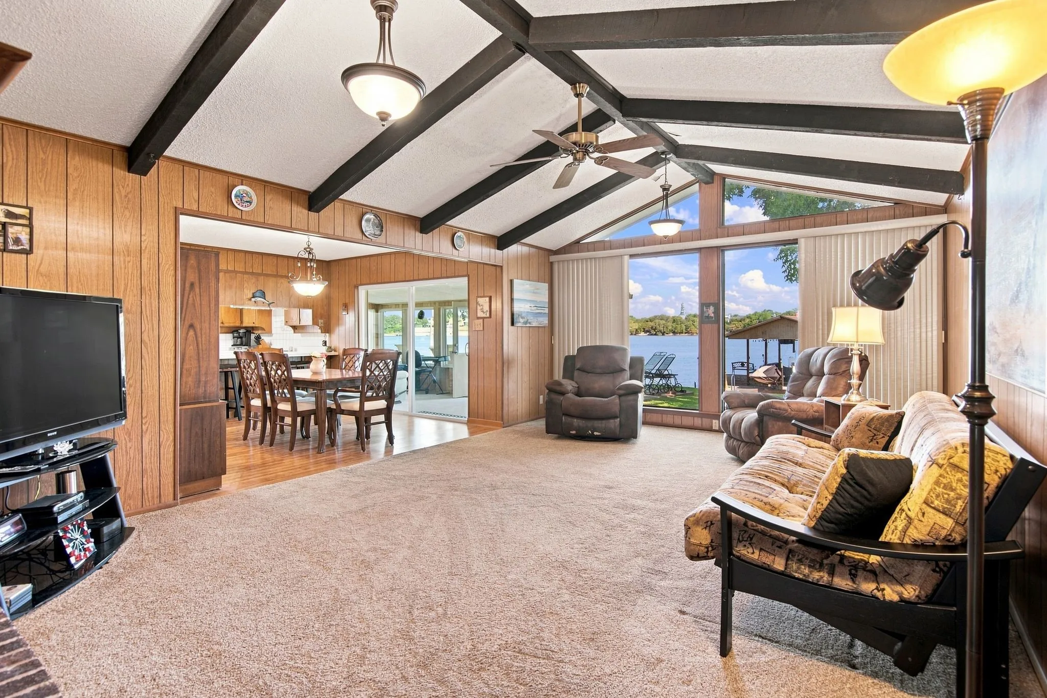 Vaulted ceiling with exposed beams, wood-paneled walls, large windows, ceiling fan, and built-in lighting fixtures