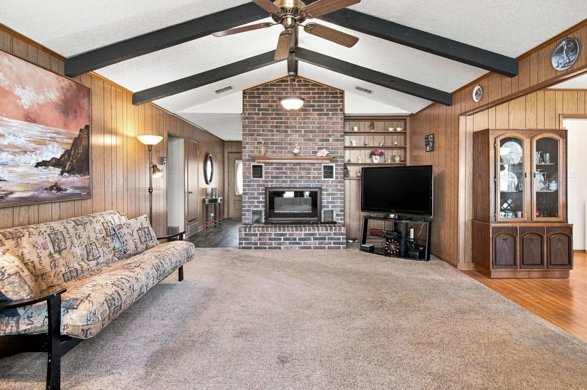 Vaulted ceilings with exposed beams, brick fireplace with built-in shelving, wood paneling, and a ceiling fan