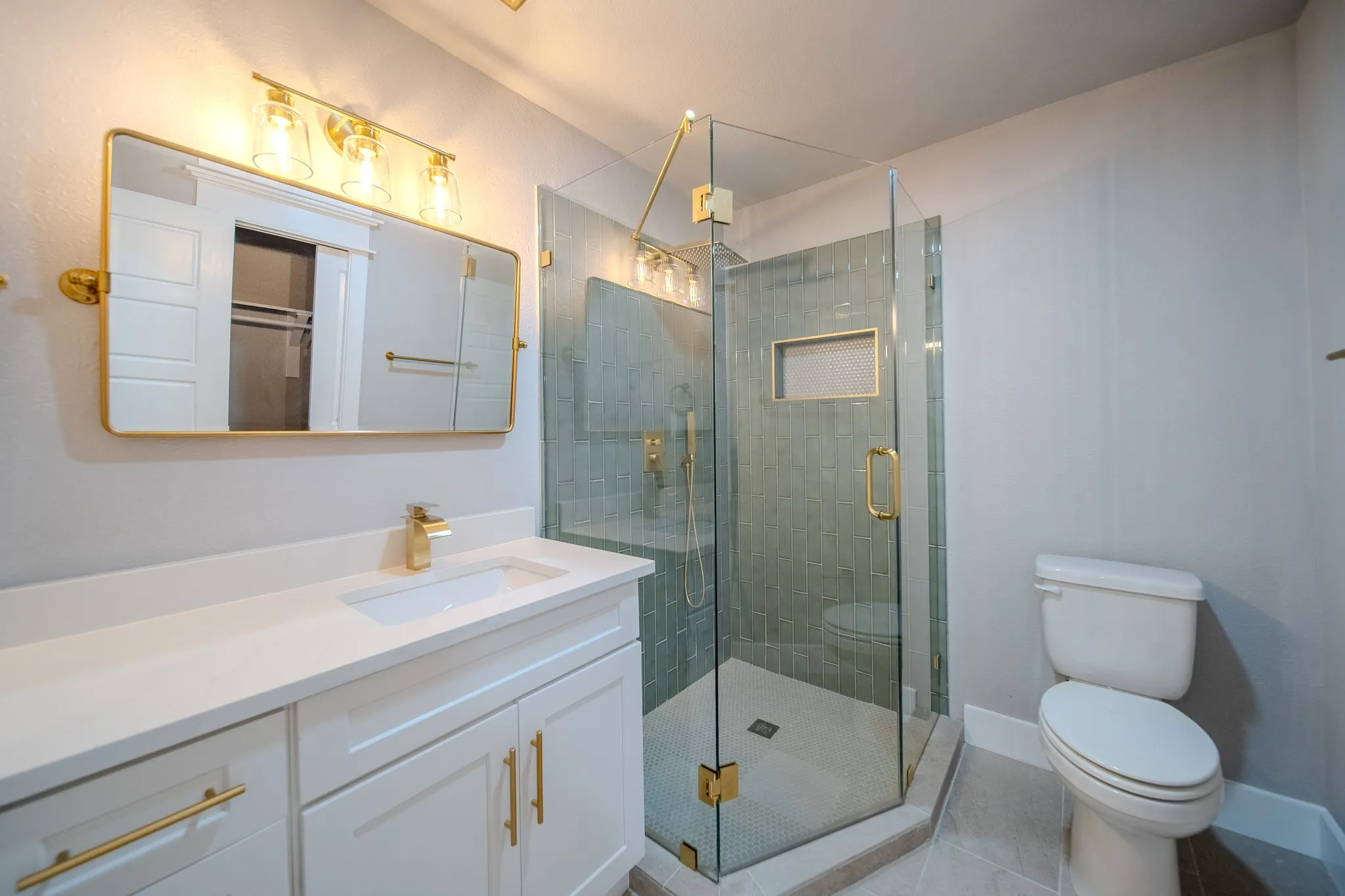 White vanity with gold-tone hardware and a rectangular undermount sink, clear glass shower enclosure with gold-tone fixtures, green subway tile shower surround, and a built-in shower niche
