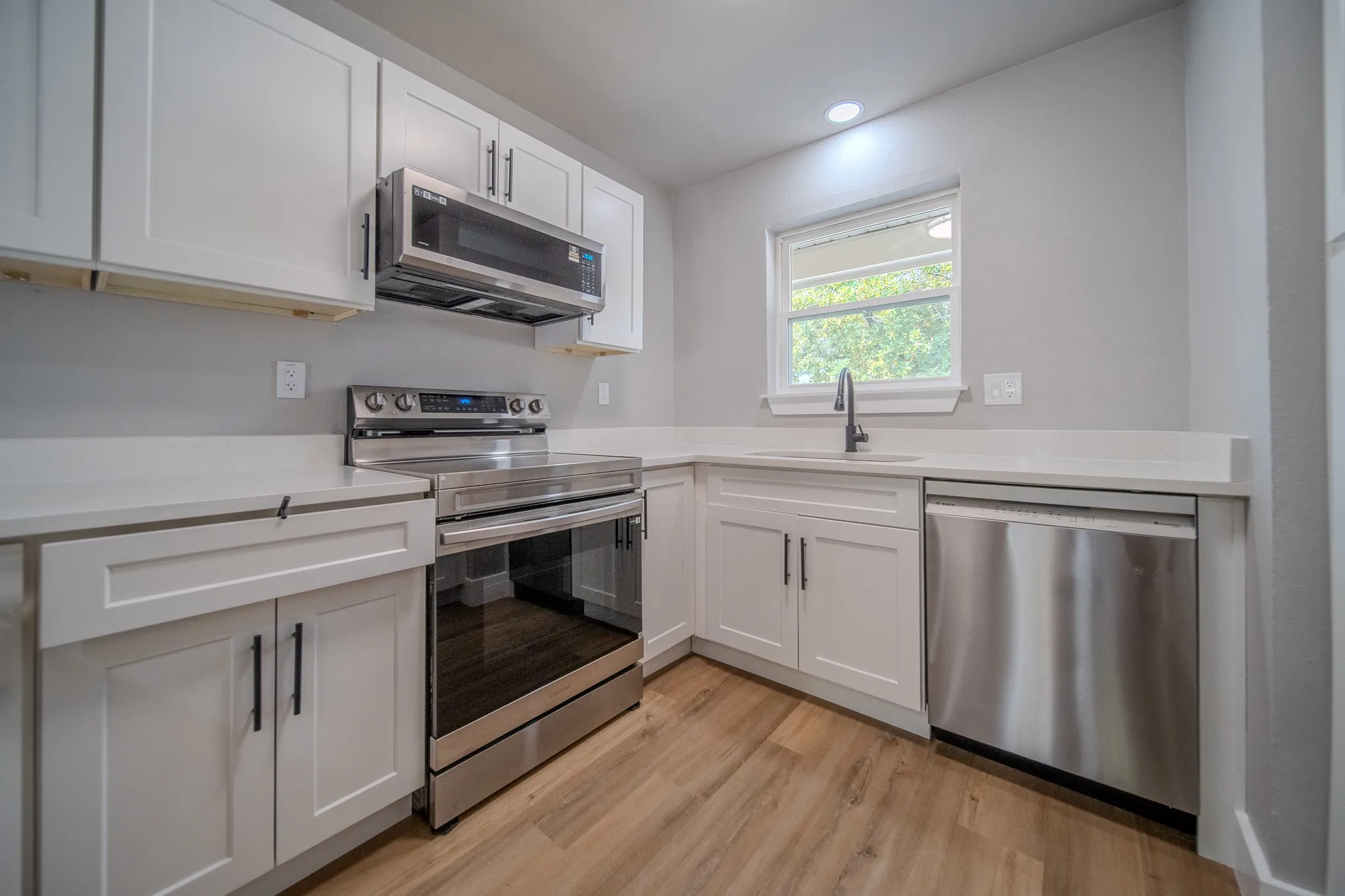 Kitchen featuring white shaker cabinetry, white countertops, wood-finish flooring, stainless steel appliances, and a window above the sink