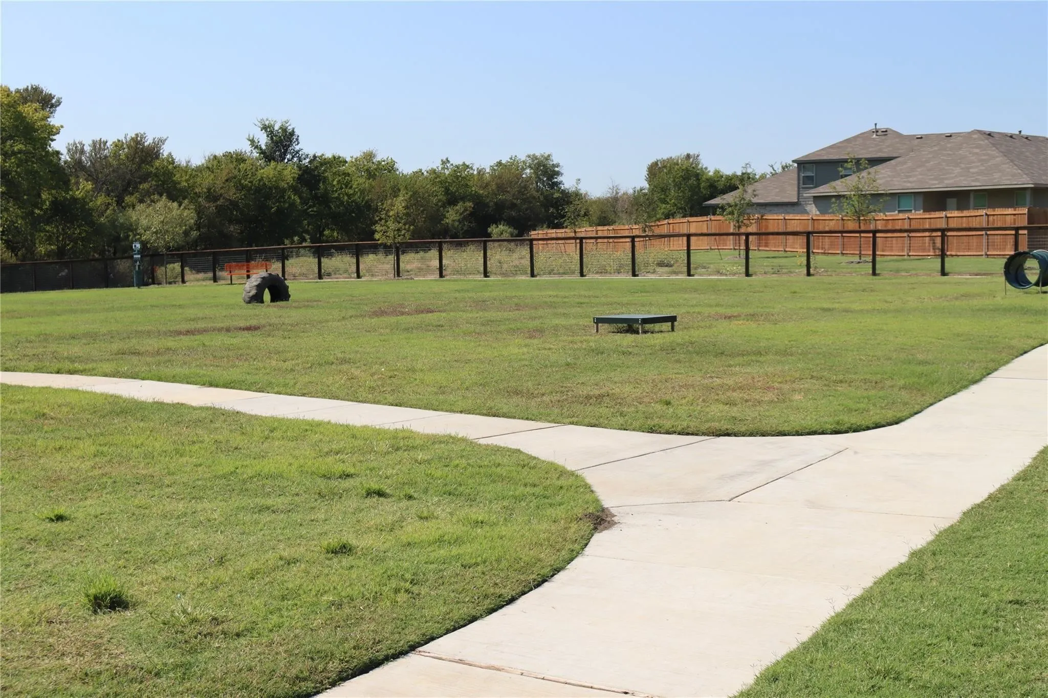 Expansive grassy area with concrete pathways, secured by a perimeter fence, and featuring various recreational elements
