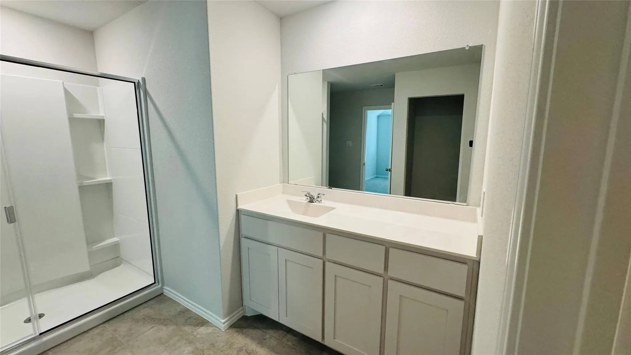 White vanity with integrated sink and chrome faucet, large frameless mirror, and glass-enclosed shower with built-in shelving