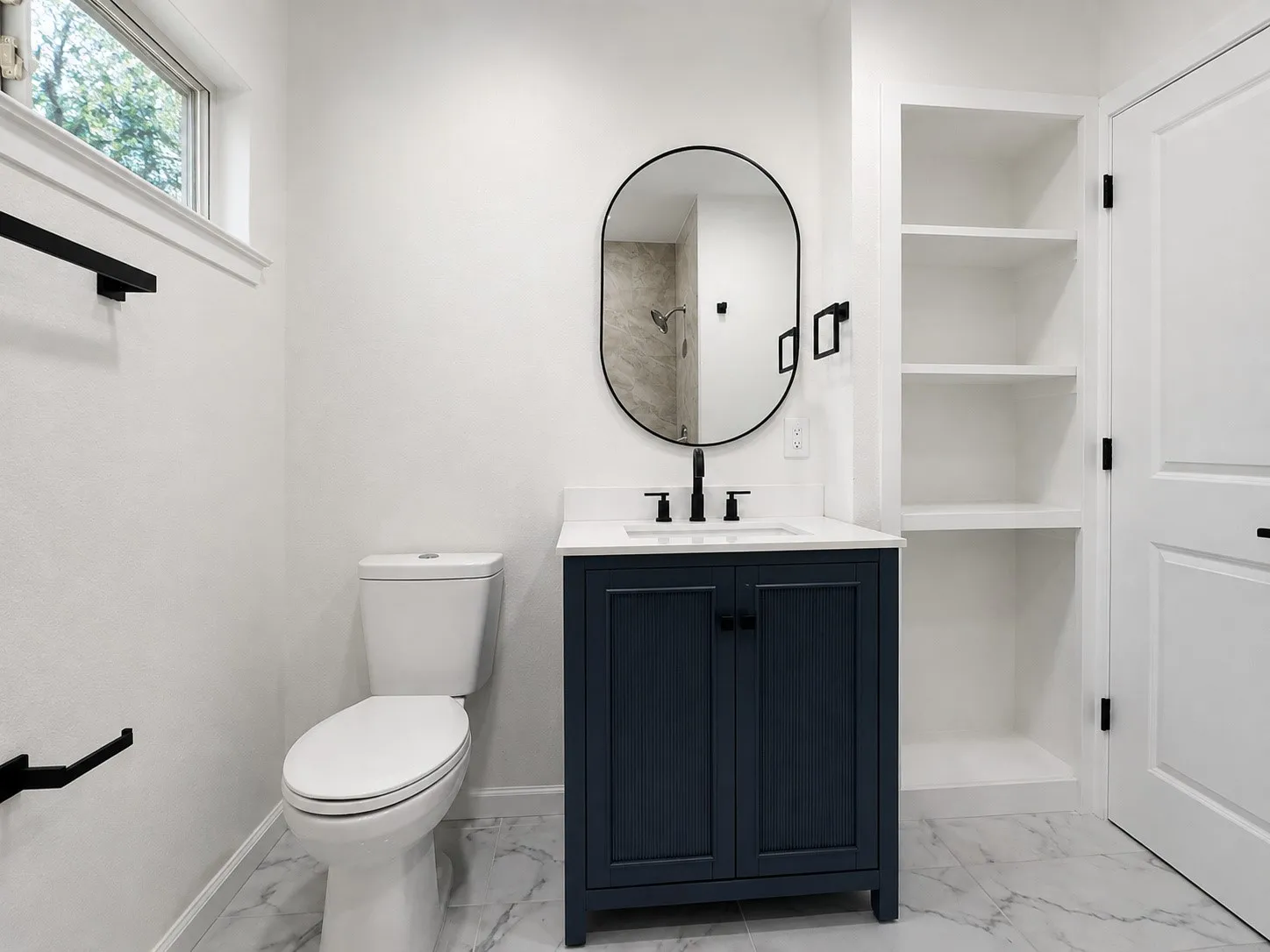 White marble-look flooring, dark blue vanity with a white countertop and undermount sink, built-in shelving, and a black-framed oval mirror