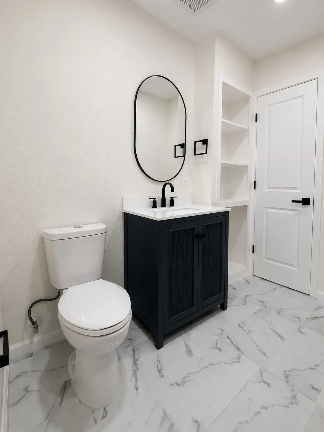 White-veined flooring, vanity with dark cabinetry, modern oval mirror, built-in shelving, and contemporary black fixtures