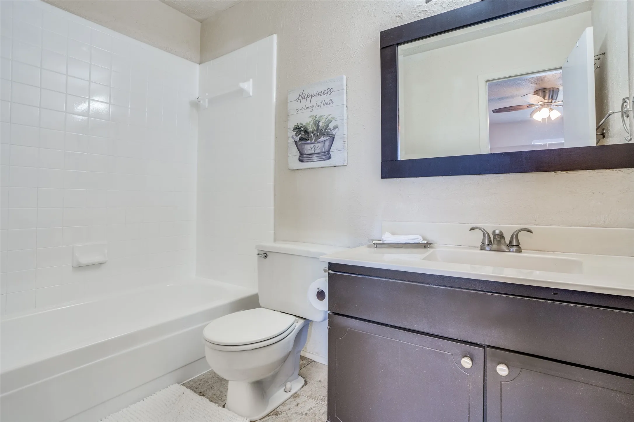 White tile surround bathtub, toilet, dark wood-finish vanity with white countertop, and framed mirror