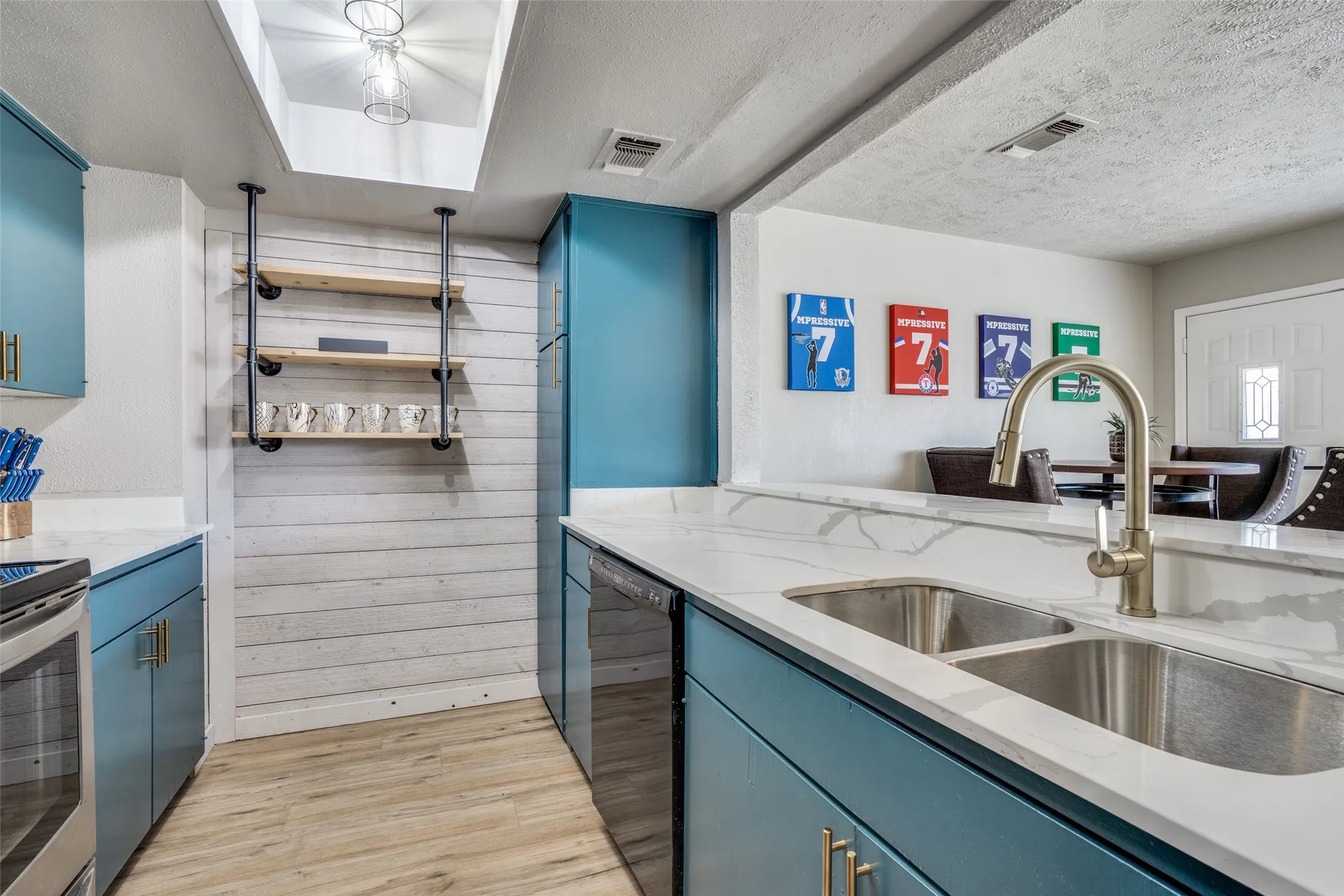 Custom blue cabinetry, stainless steel double basin sink, gold-tone gooseneck faucet, light-toned wood-finish flooring, and white shiplap accent wall