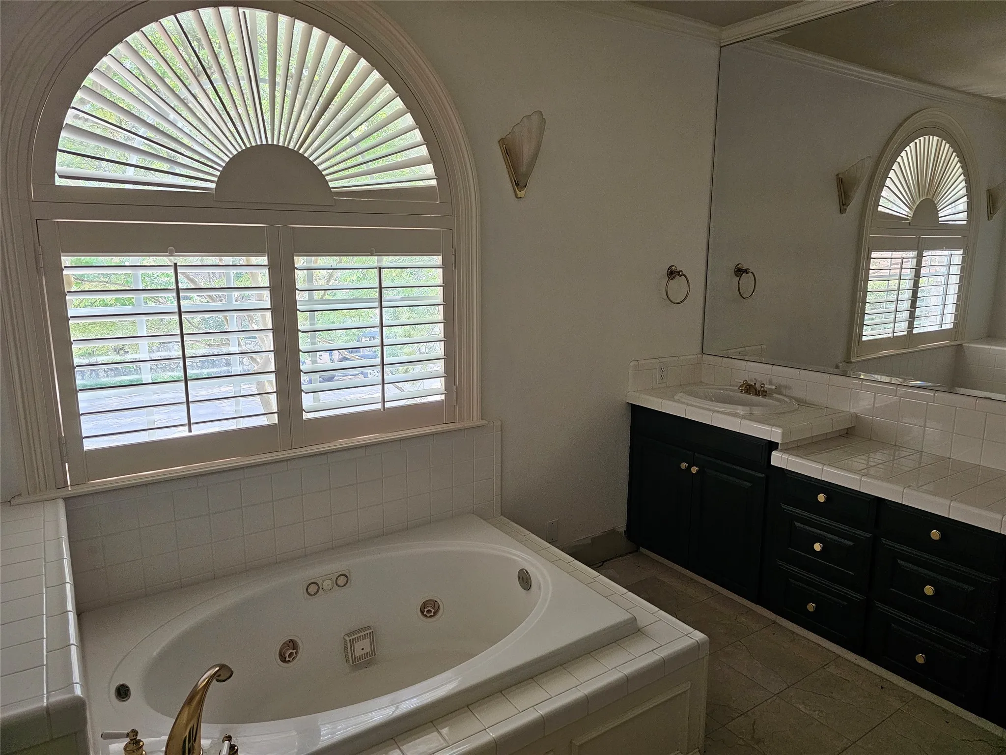 Arched window with plantation shutters, built-in jetted tub with tile surround, vanity with dark wood-finish cabinetry, integrated sink, and large wall-mounted mirror