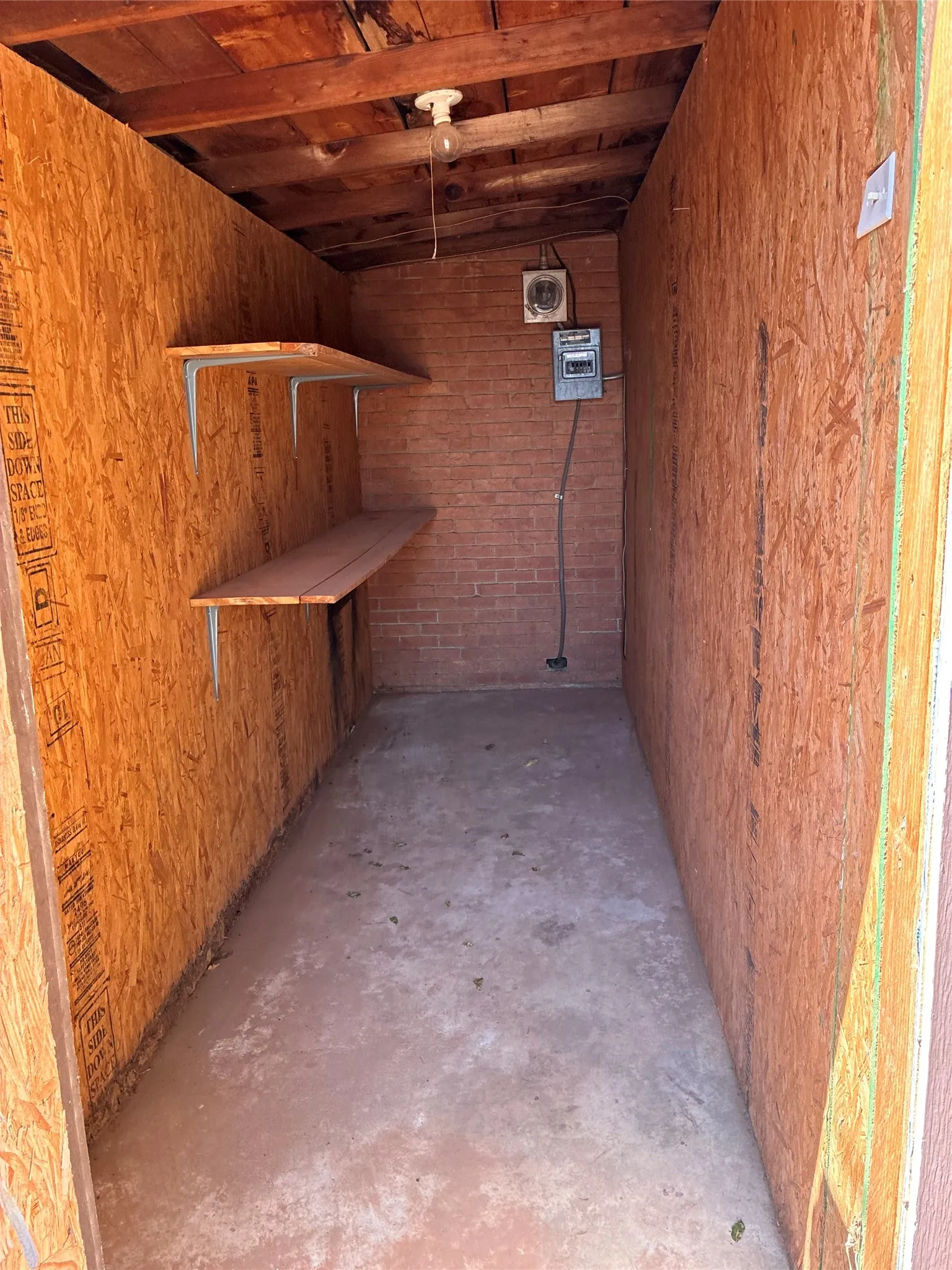 Storage space featuring a concrete floor, built-in shelving, brick accent wall, and exposed wood ceiling joists