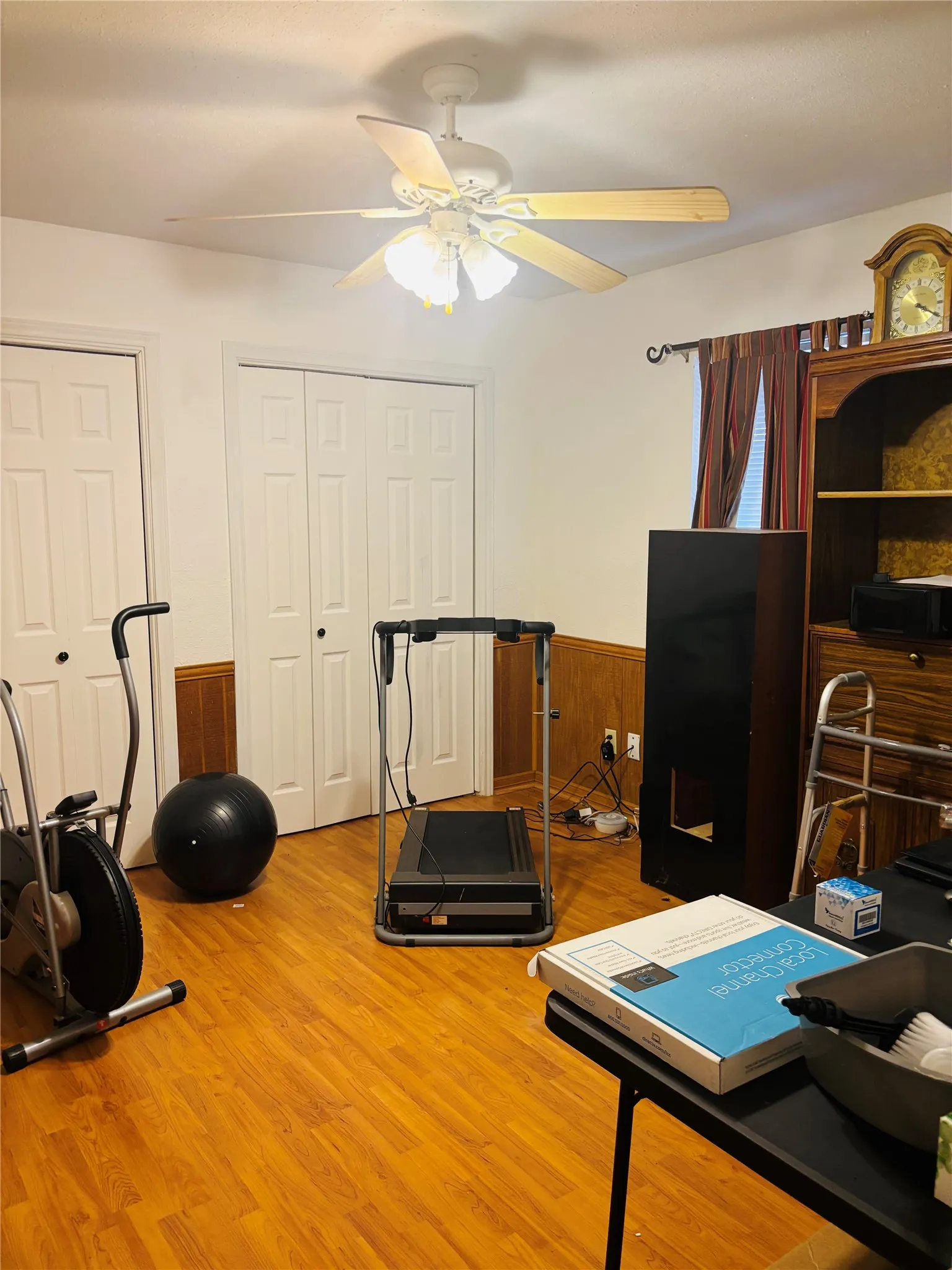 Wood-finish flooring, two panel doors, wainscoting, and a ceiling fan with integrated lighting