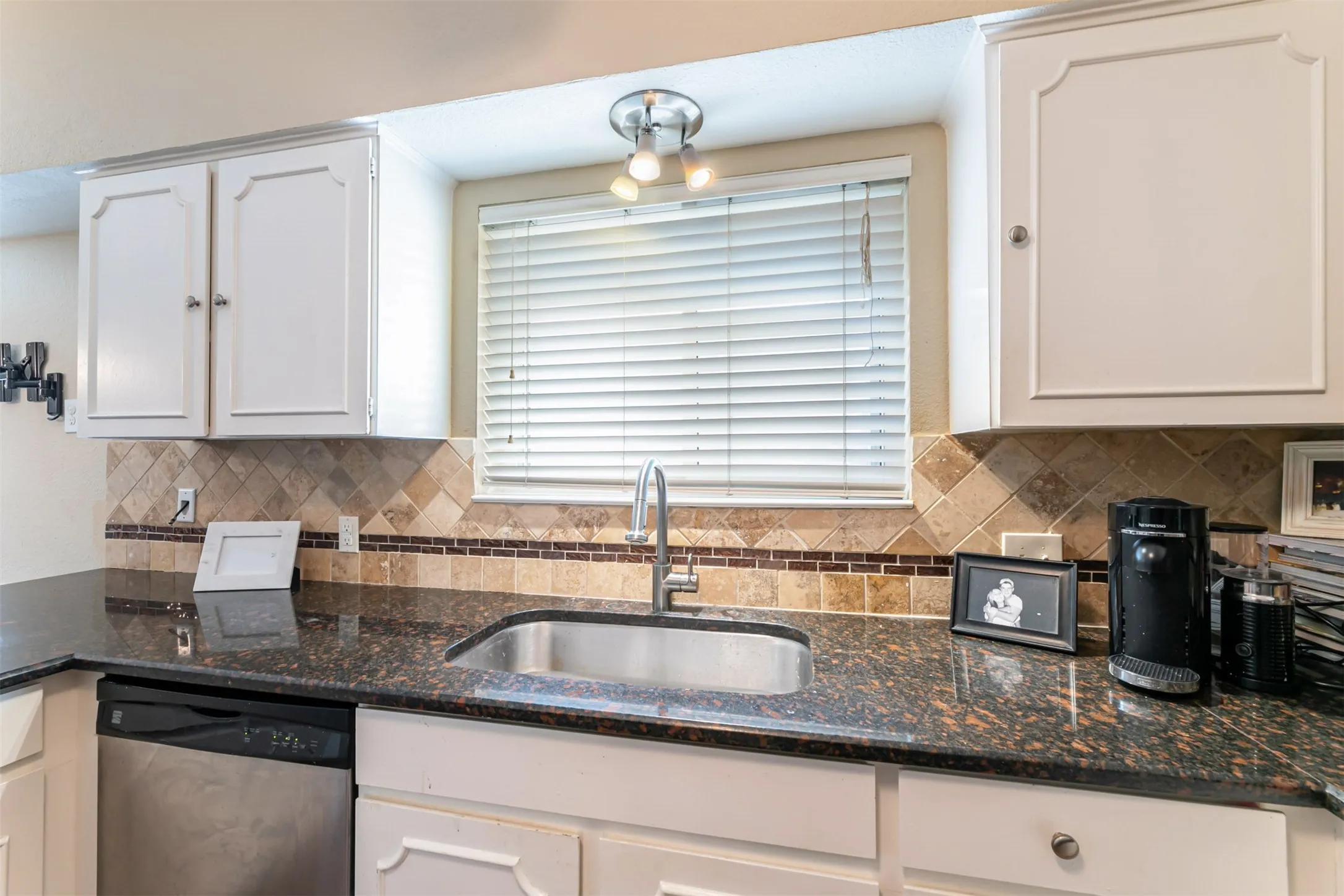 Dark granite countertops, stainless steel sink with a high-arc faucet, tiled backsplash with decorative border, white cabinetry with panel detailing, and a stainless steel dishwasher