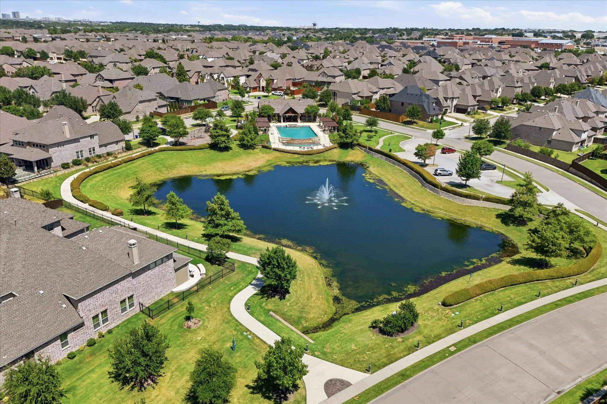 Community pond with a decorative fountain, surrounded by paved walking paths and manicured landscaping, featuring a resort-style swimming pool, and a community building with a covered patio