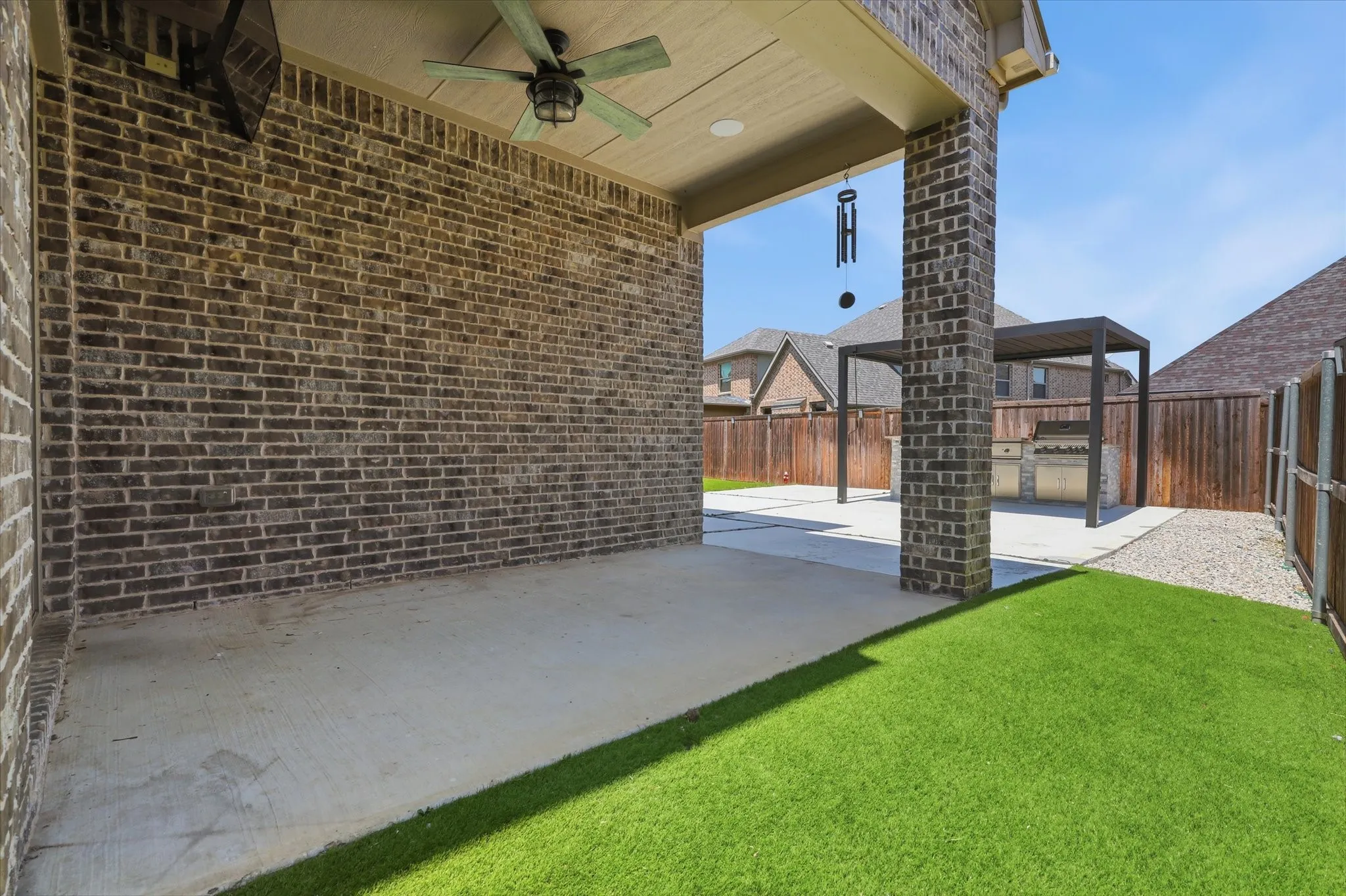Covered patio with a ceiling fan, brick exterior, concrete paving, artificial turf, and a built-in outdoor kitchen with a pergola