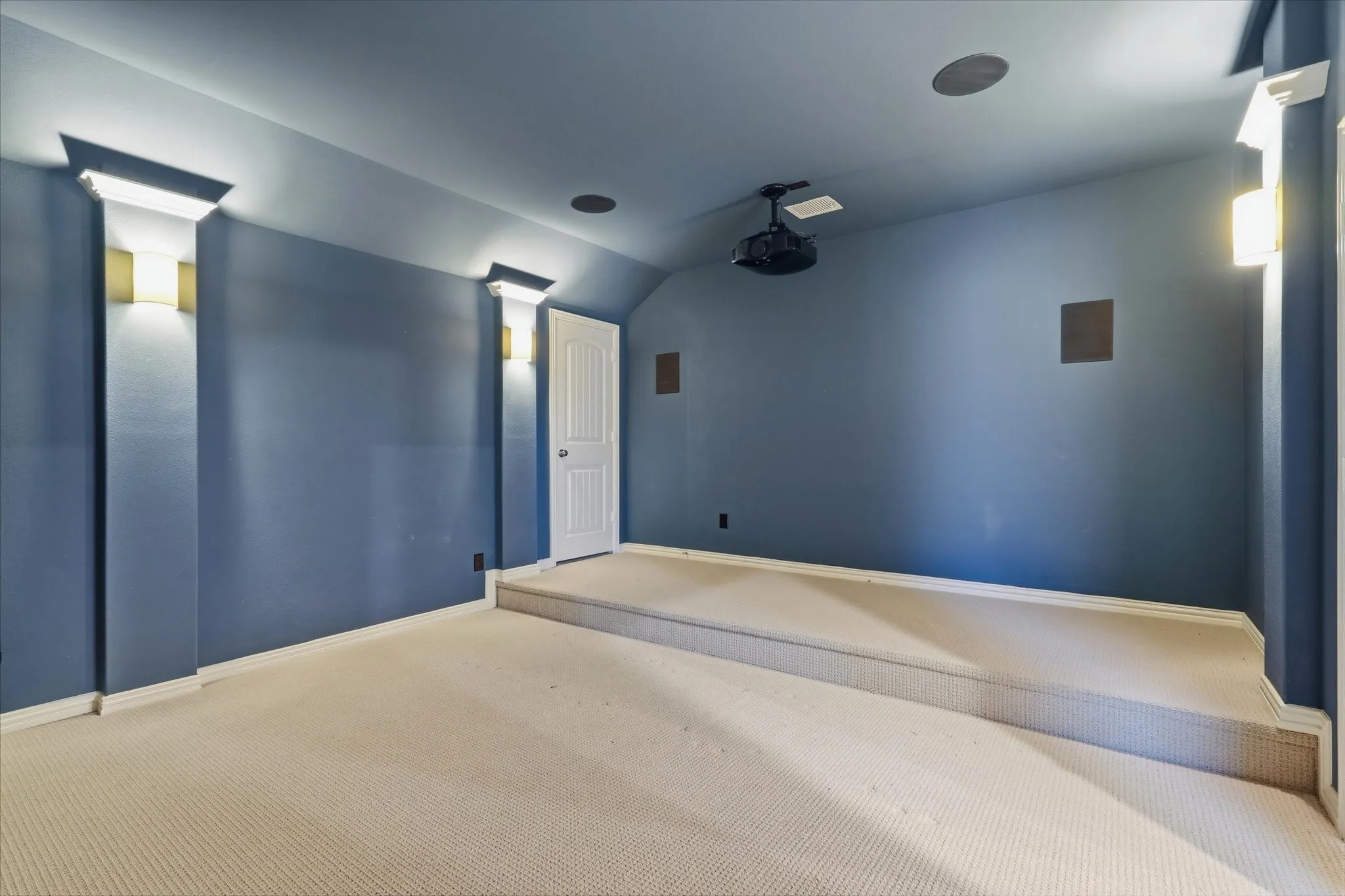 Carpeted room featuring a raised platform, integrated wall speakers, ceiling-mounted projector, and decorative wall sconces