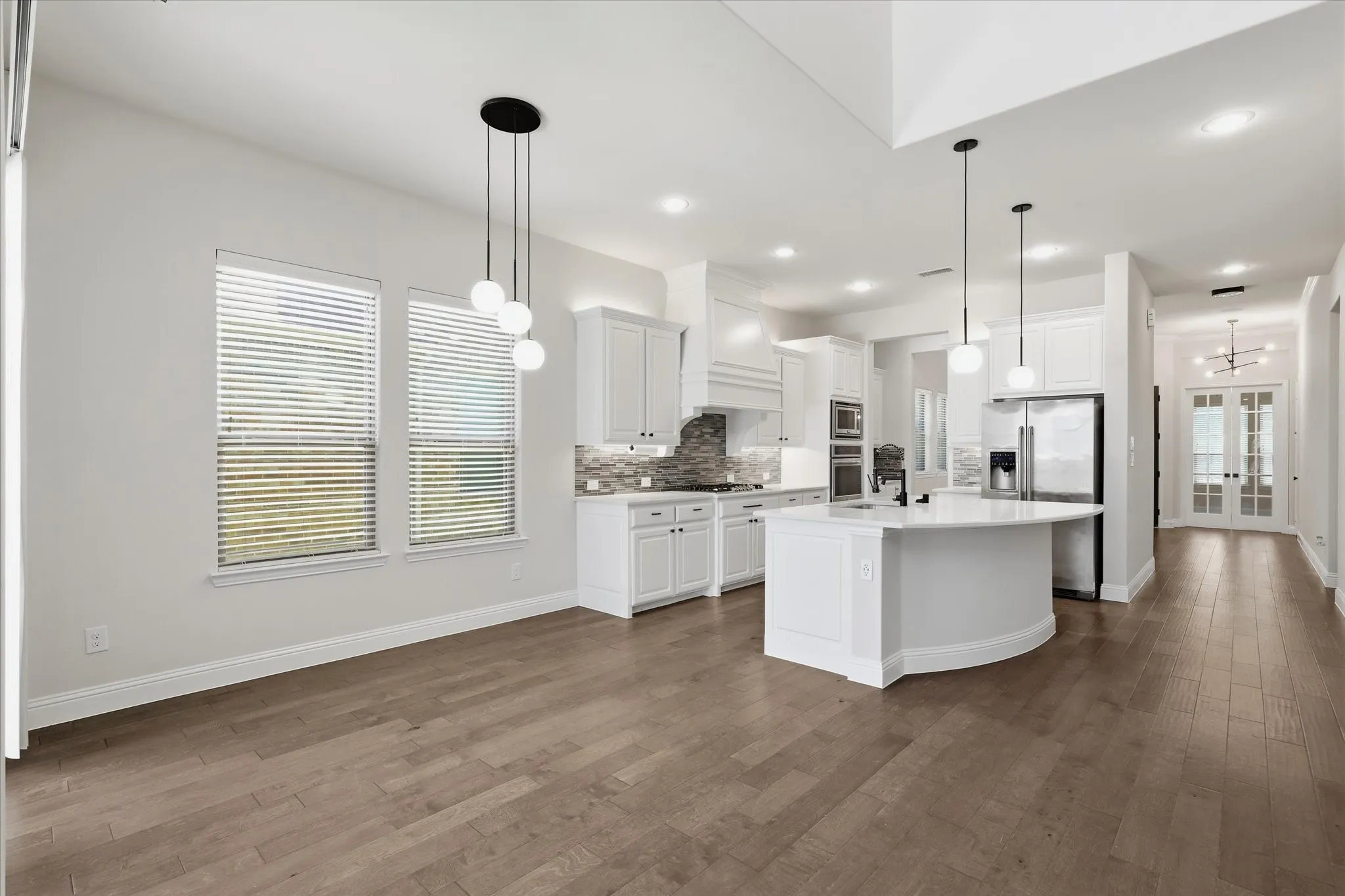 Open-concept floor plan featuring wood flooring, a kitchen with a curved island, white cabinetry, and stainless steel appliances, pendant lighting  and breakfast area.