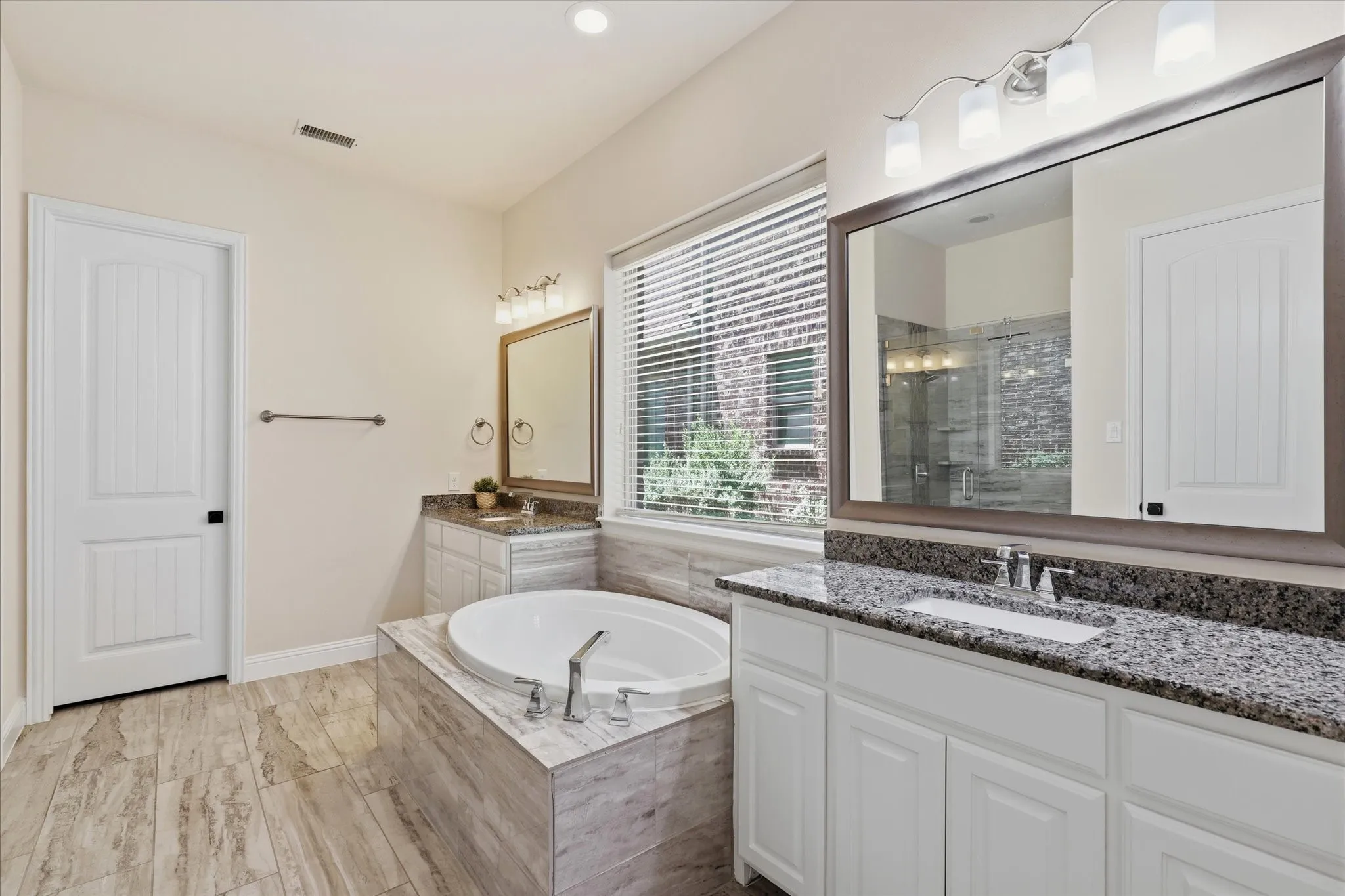 Built-in soaking tub, dual vanities with granite countertops, wood flooring, and a large window with blinds