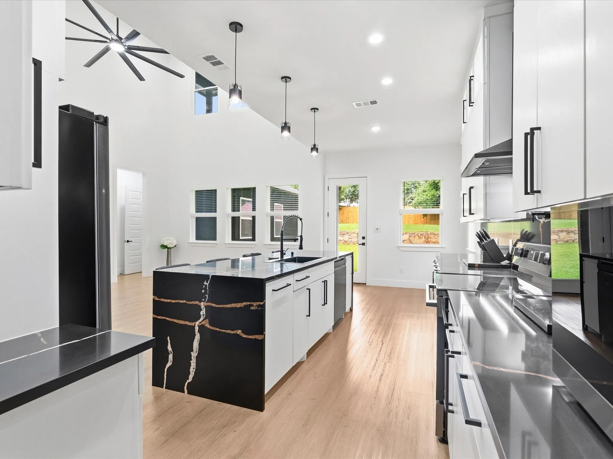 Modern kitchen featuring a waterfall island with a black and gold finish, light wood-finish flooring, white cabinetry with black hardware, and black countertops