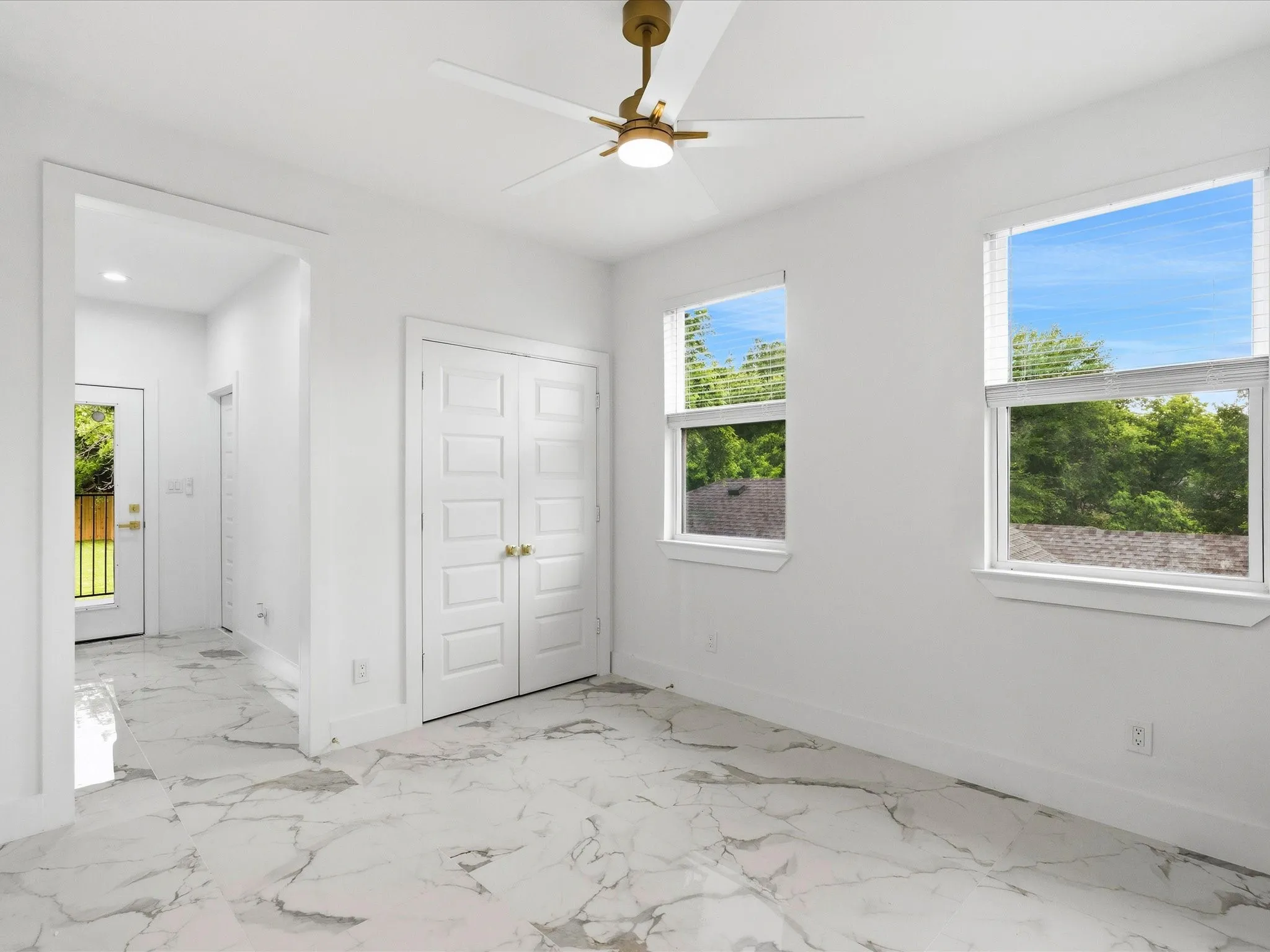 White interior walls, two windows, gold-toned ceiling fan, double closet doors, and marble-look tile flooring
