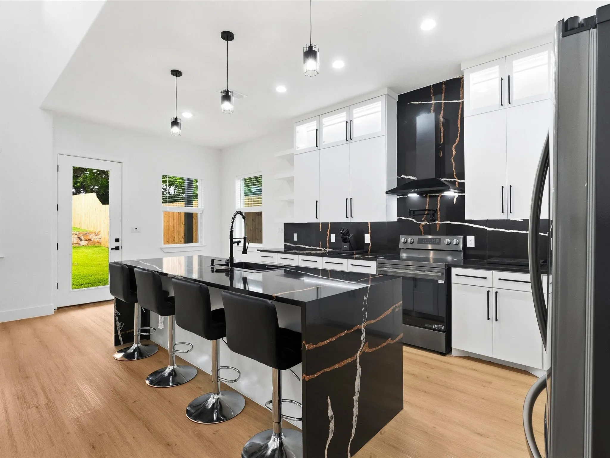 Contemporary kitchen featuring a large island with integrated sink, black countertops with distinctive veining, white cabinetry, stainless steel appliances, and light wood-finish flooring