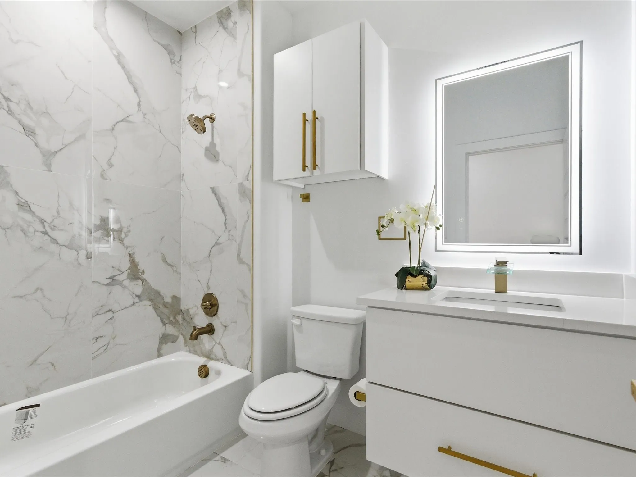Wall-mounted vanity with integrated sink, white cabinetry with gold-tone hardware, backlit rectangular mirror, full bathtub with gold-tone fixtures, and marble-patterned wall tiling