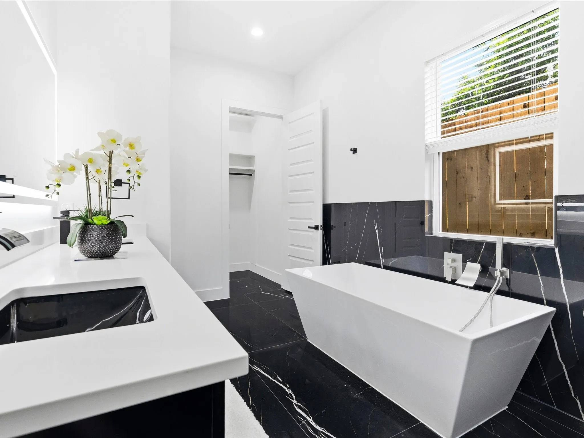 Black and white marbled flooring, freestanding soaking tub, built-in vanity with integrated sink, black tiled wall, and natural light from a large window
