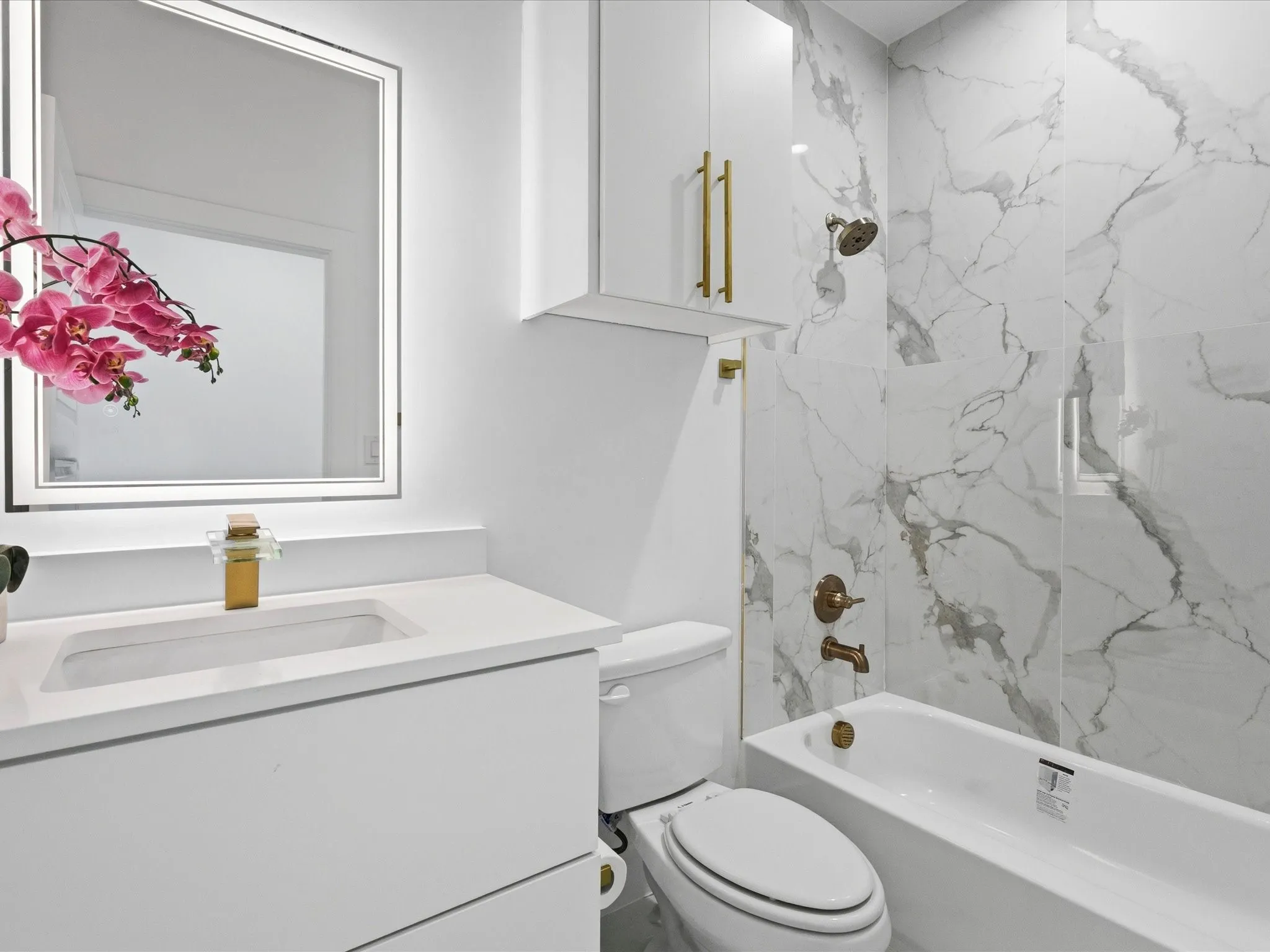 Floating vanity with an integrated sink and gold-tone faucet, illuminated mirror, and a built-in bathtub with a shower featuring large format veined wall tiles and matching gold-tone fixtures