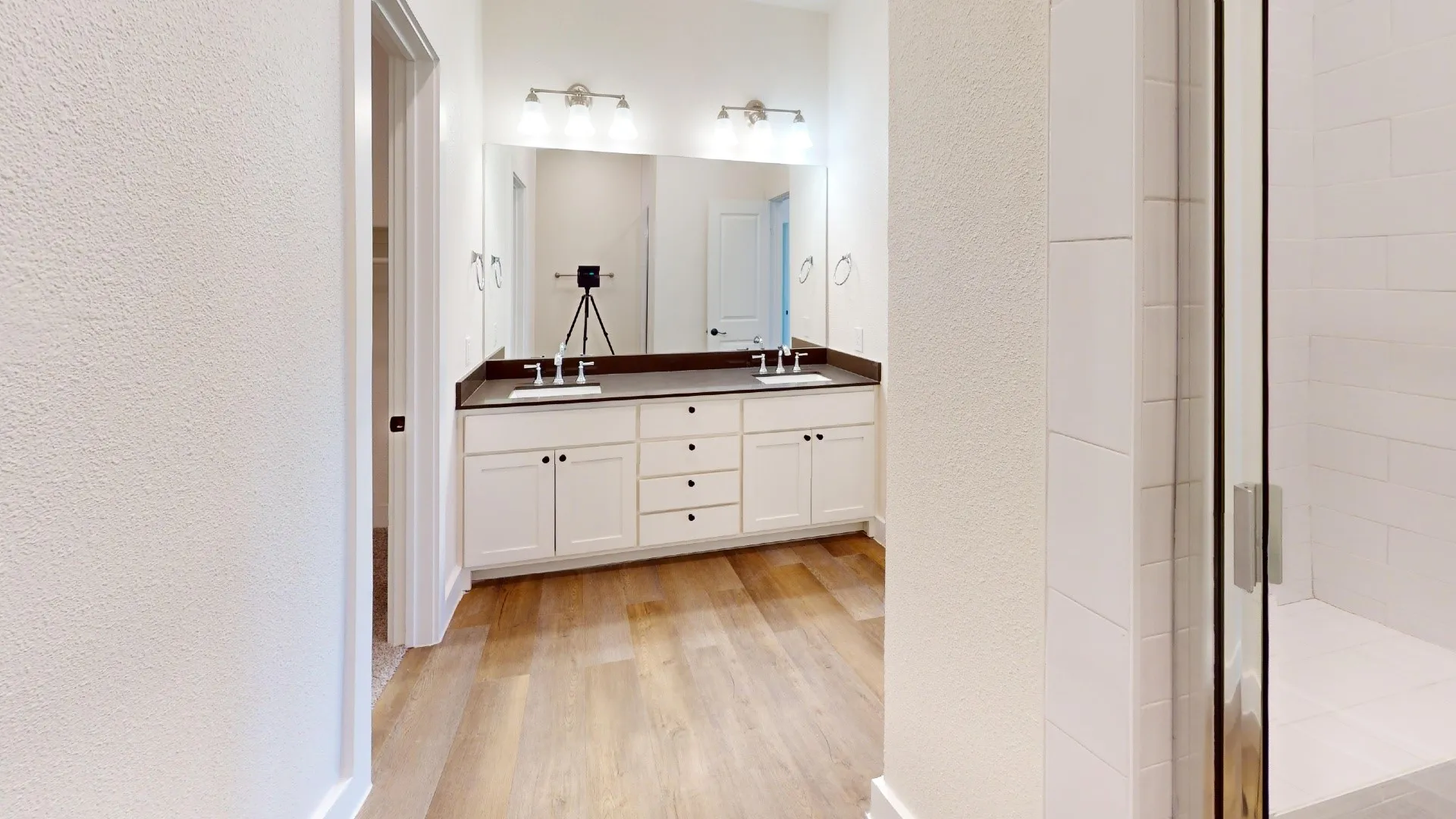 Dual vanity with white cabinetry and dark countertops, wood-finish flooring, and a glass-enclosed shower