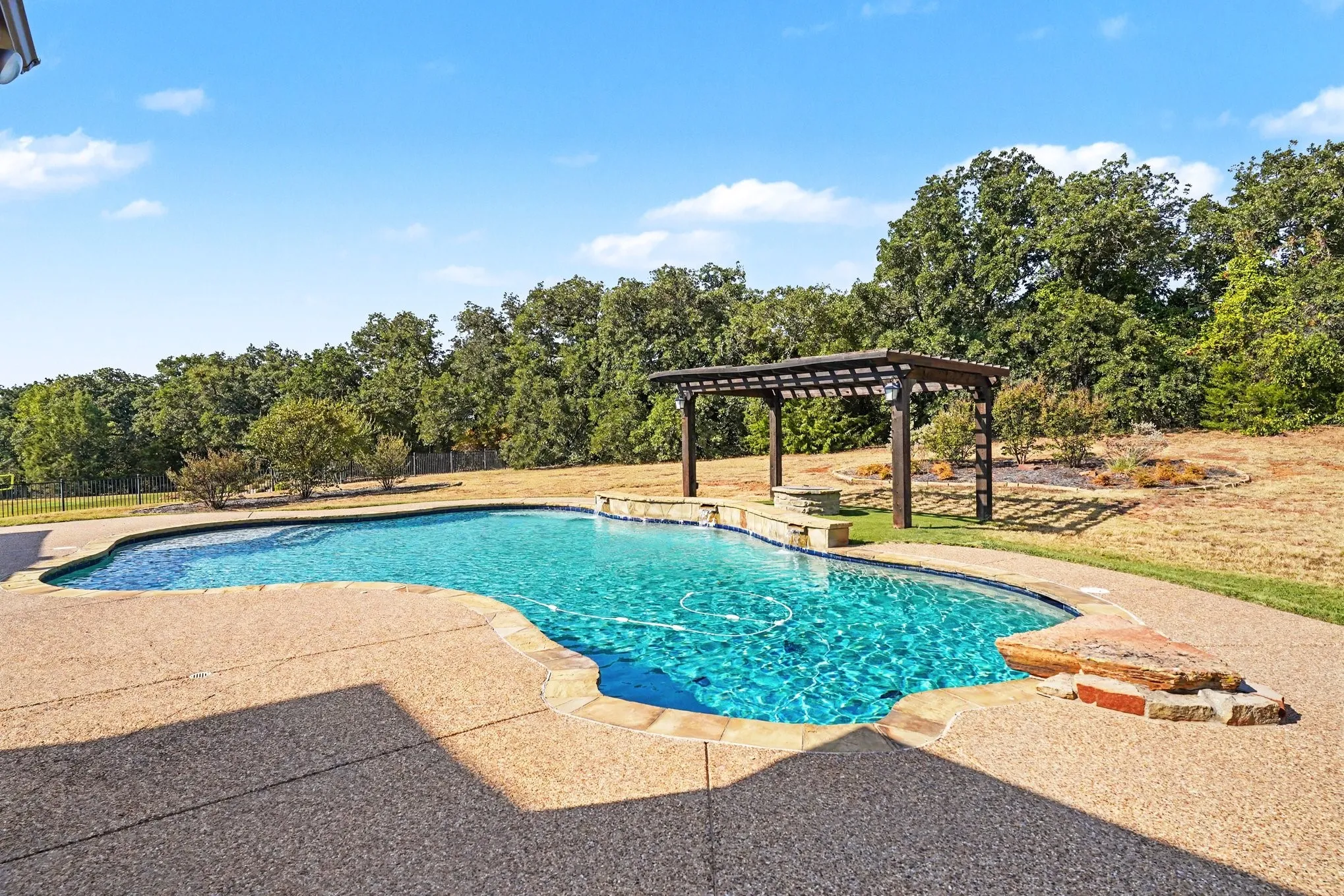 Curved swimming pool with an integrated water feature, surrounded by a textured concrete patio