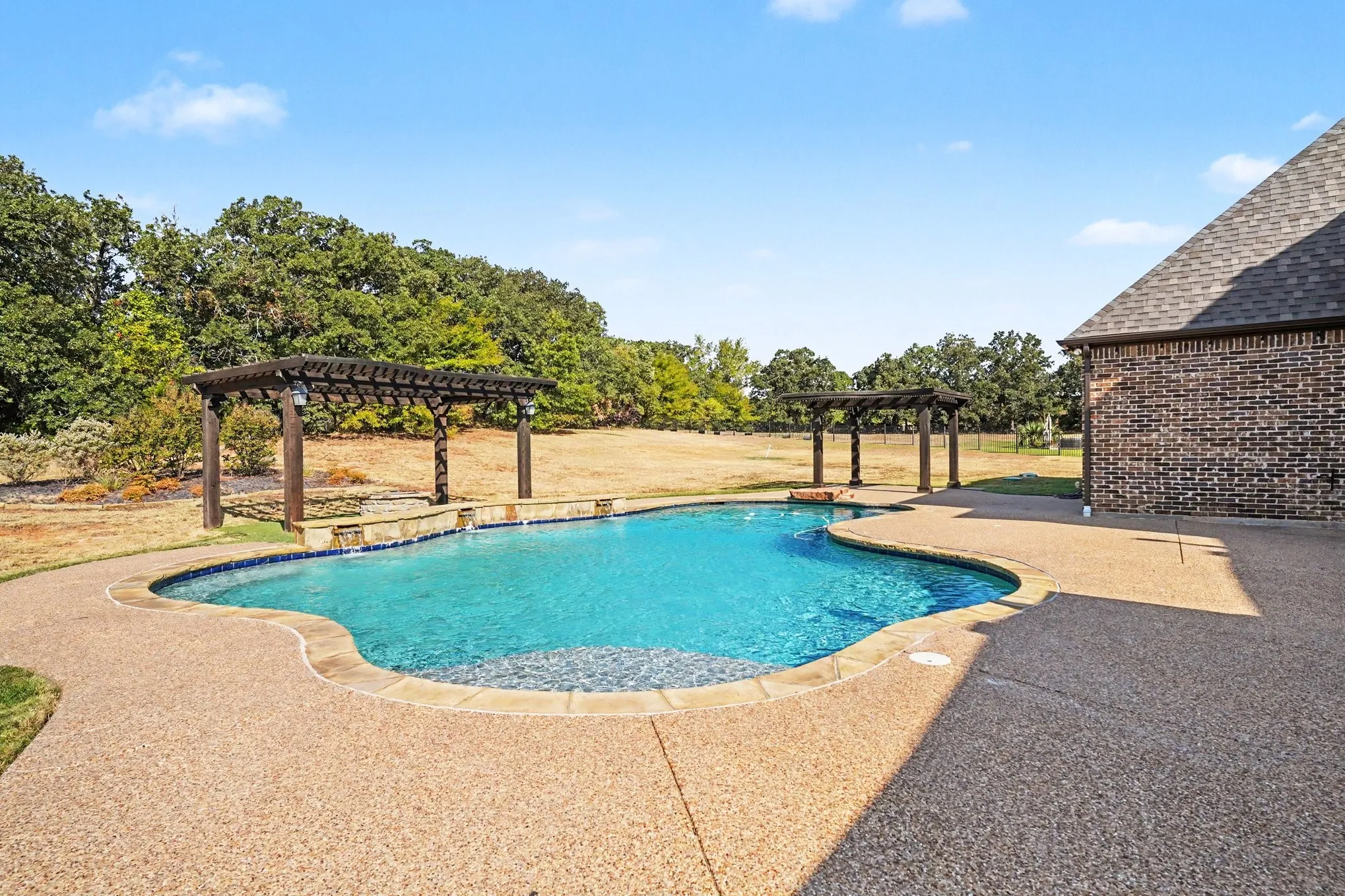 Freeform swimming pool with a surrounding aggregate concrete patio, two dark wood pergolas, and a brick exterior wall